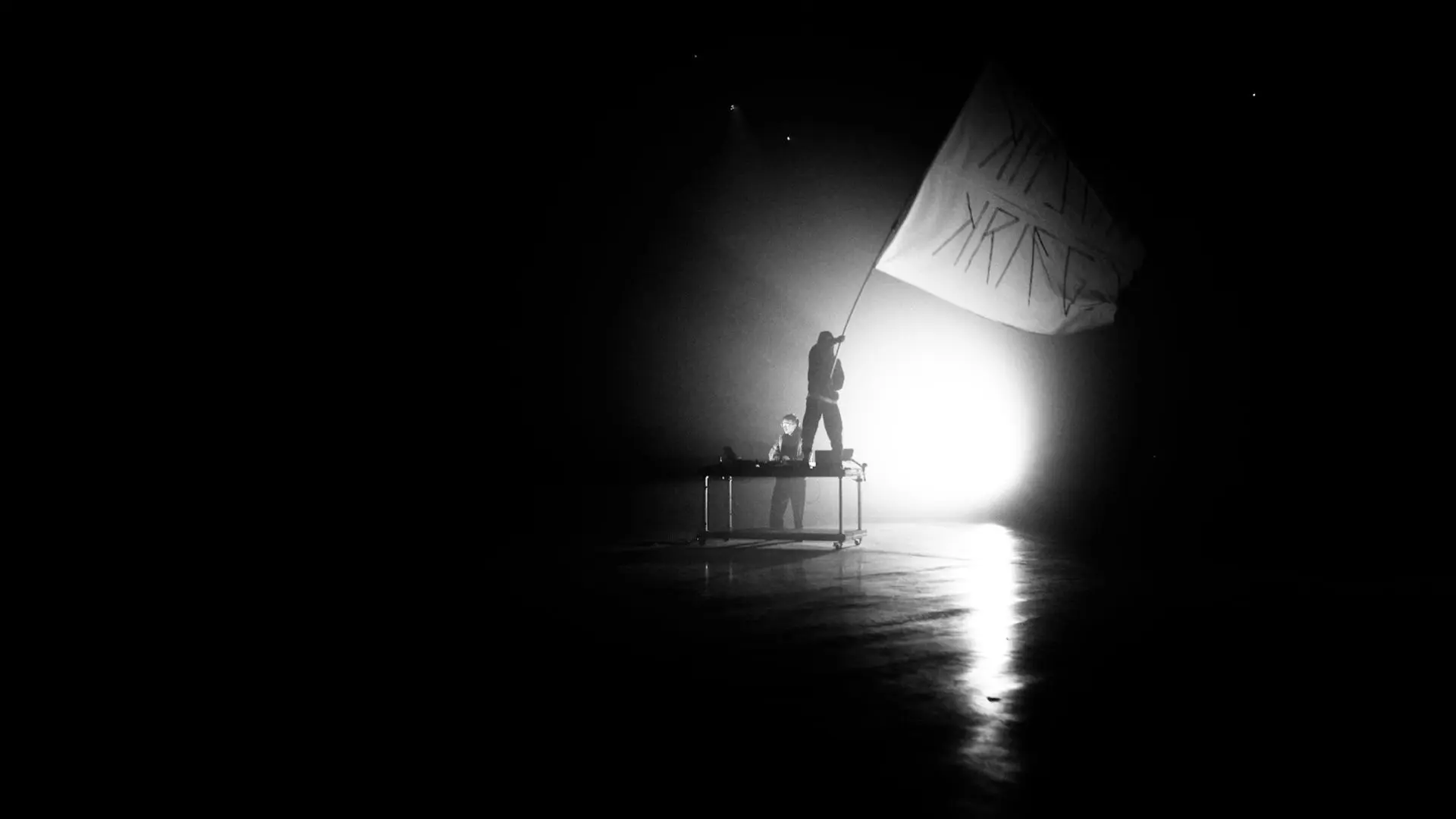Silhouette einer Person, die auf einem Tisch steht und eine große Fahne hält, im Gegenlicht mit starkem Lichtschein im Hintergrund