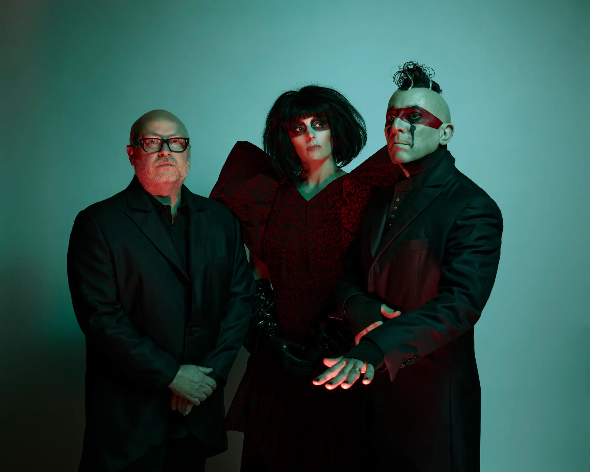 Drie leden van Puscifer poseren tegen een groenblauwe achtergrond. Links staat Mat Mitchell met bril in zwart pak, in het midden Carina Round met zwarte pruik en opvallende outfit, rechts Maynard James Keenan met hanenkam en rode gezichtsverf.
