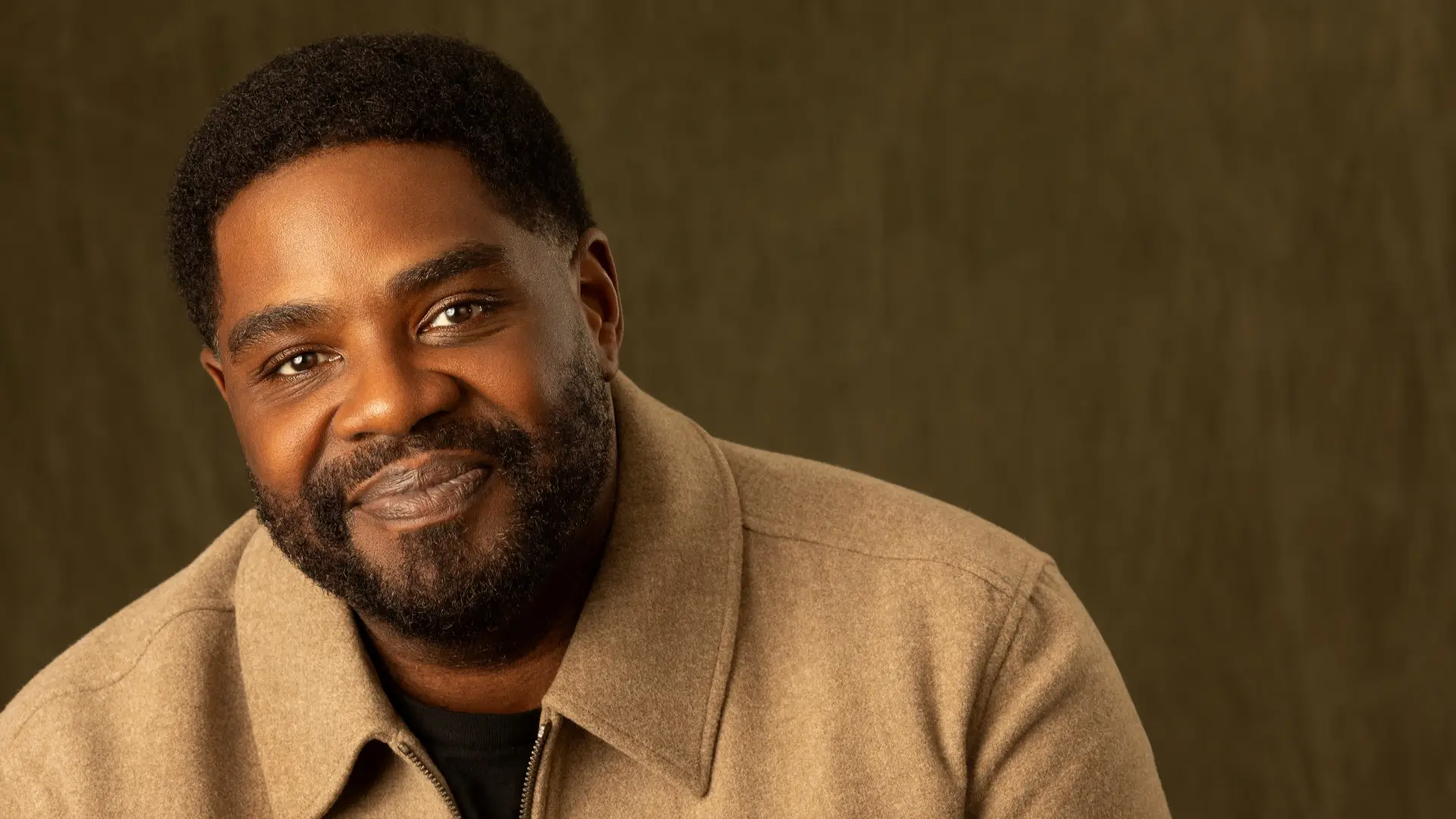 Portret van Ron Funches, glimlachend, met kort haar en baard, gekleed in een lichtbruine jas tegen een warme, onscherpe achtergrond