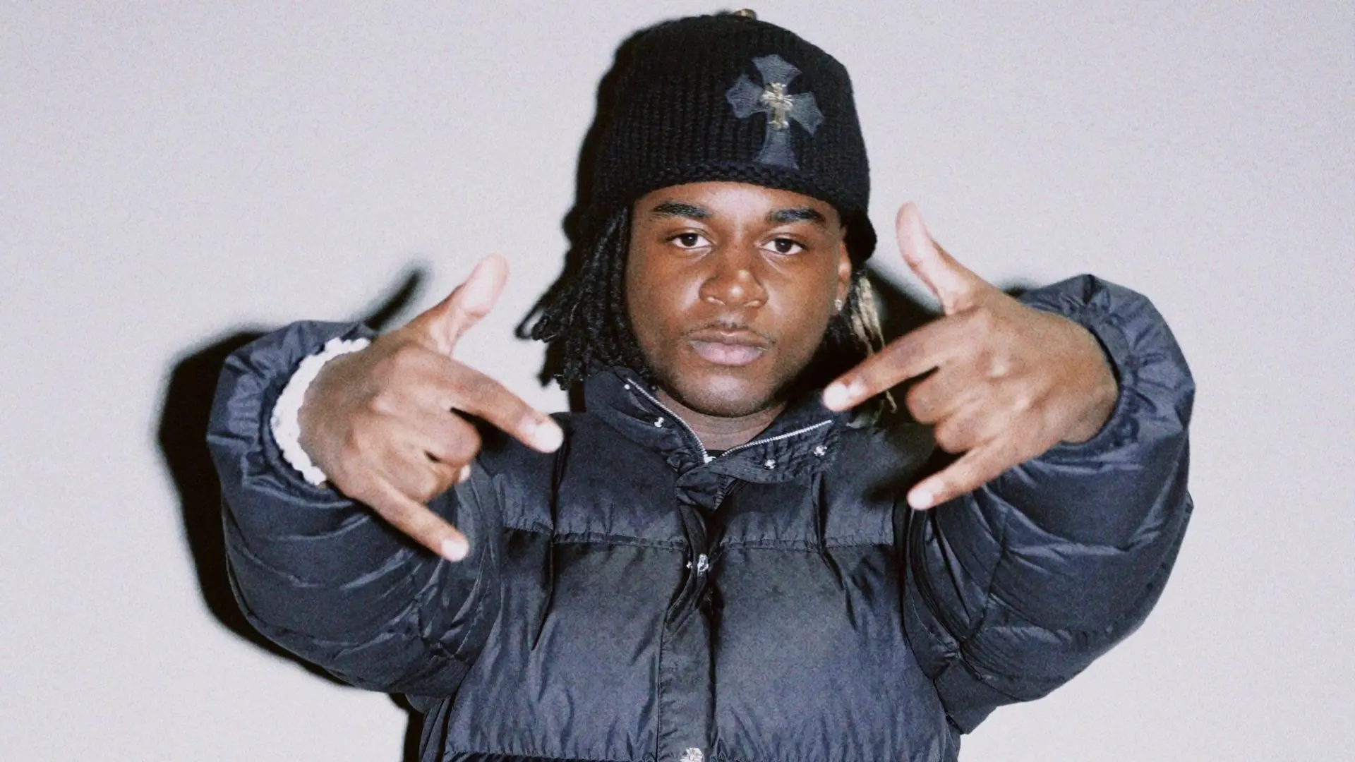 Ken Carson in zwarte gewatteerde jas en zwarte muts die met beide handen handgebaren naar de camera maakt.