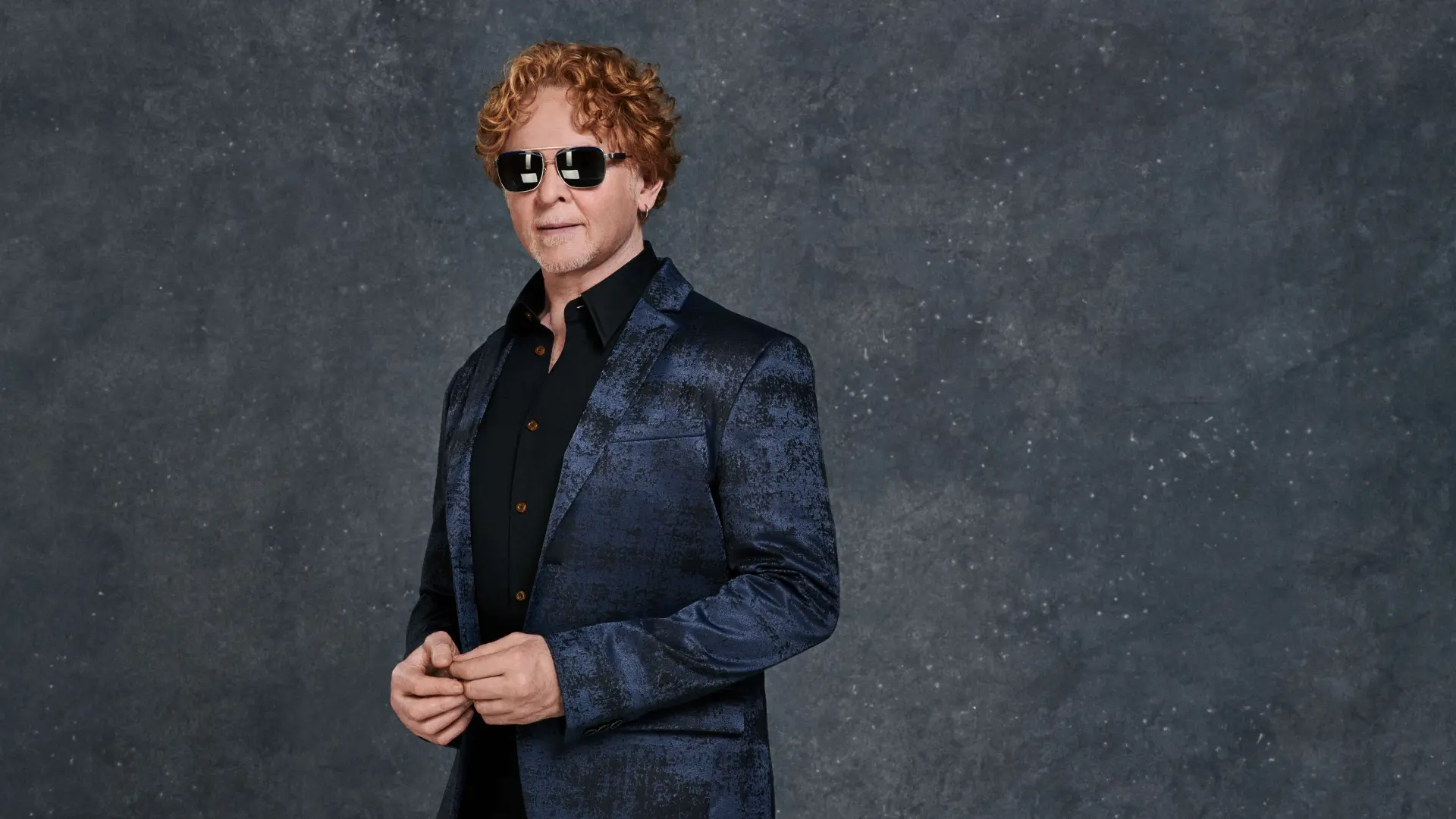 Simply Red-zanger Mick Hucknall in donkerblauw colbert en zonnebril tegen een donkergrijze achtergrond.