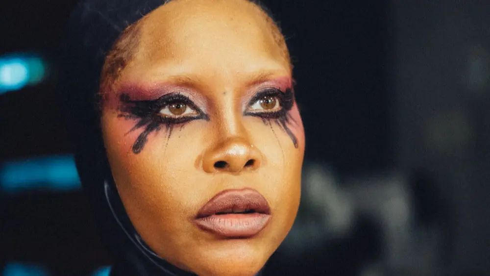 Erykah Badu draagt een opvallende gestreepte outfit en theatrale oogmake-up terwijl ze recht vooruit kijkt in een close-upportret.