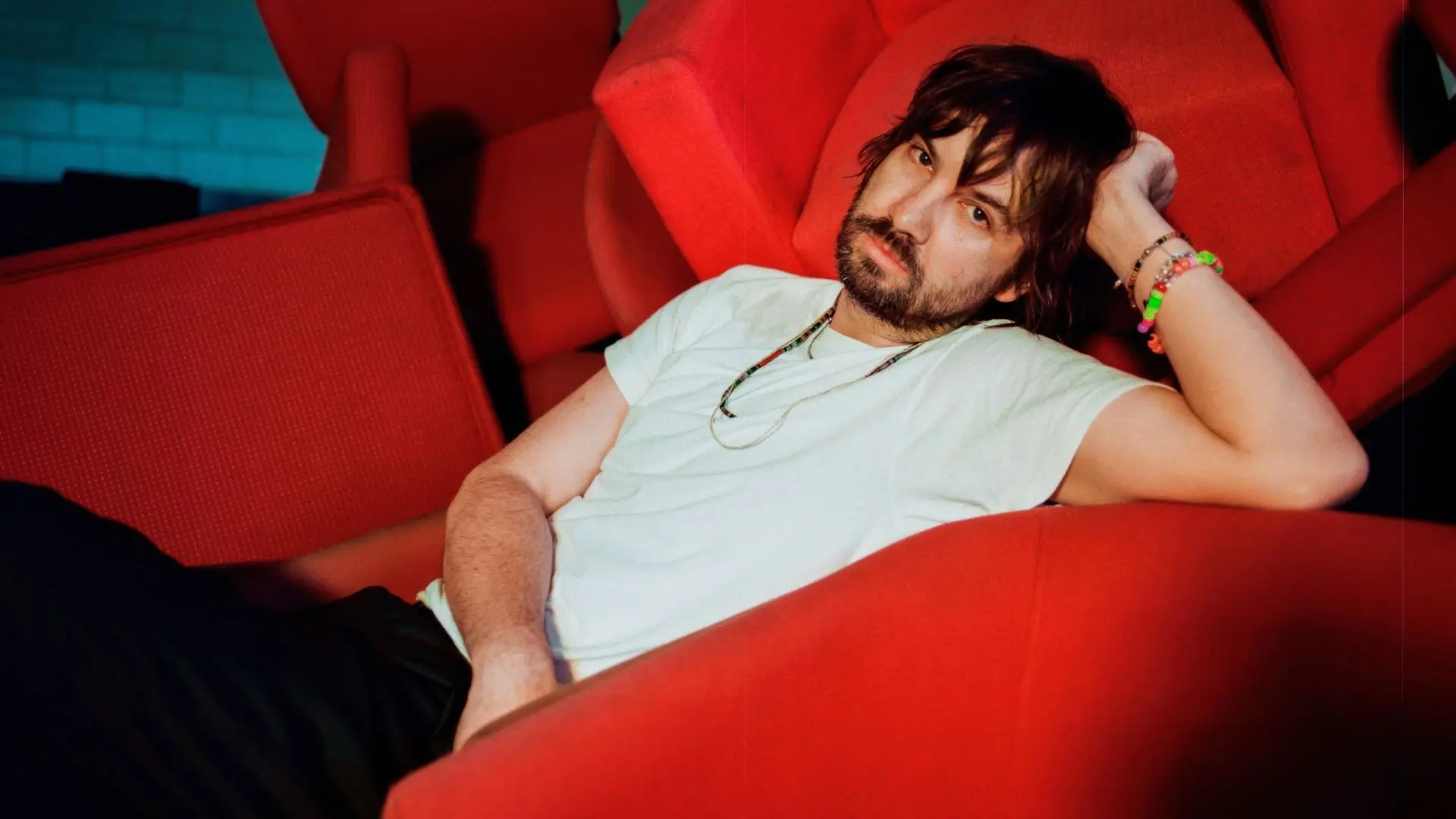 Tame Impala met halflang donker haar en baard ligt ontspannen in een rode fauteuil, draagt een licht T-shirt en kleurrijke armbanden; op de achtergrond meerdere rode stoelen.