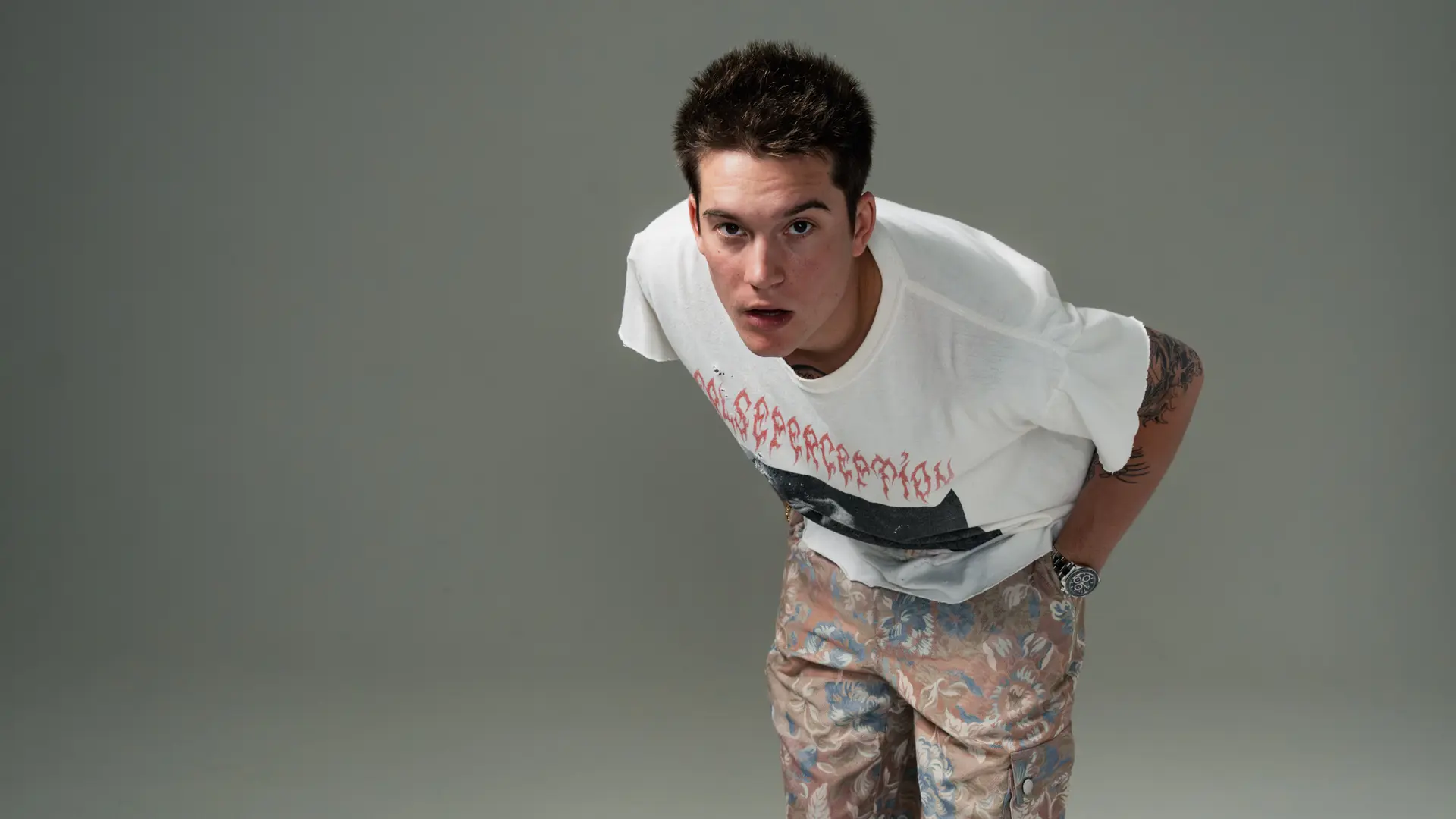 Cameron Whitcomb leunt voorover naar de camera in een wit T-shirt en gebloemde broek, met zichtbare armtattoos tegen een grijze achtergrond.