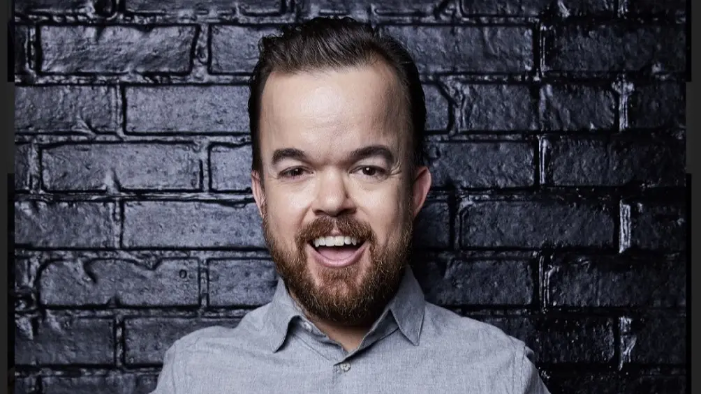 Portret van Brad Williams met een lach, voor een zwarte bakstenen muur, gekleed in een lichtgrijs overhemd.