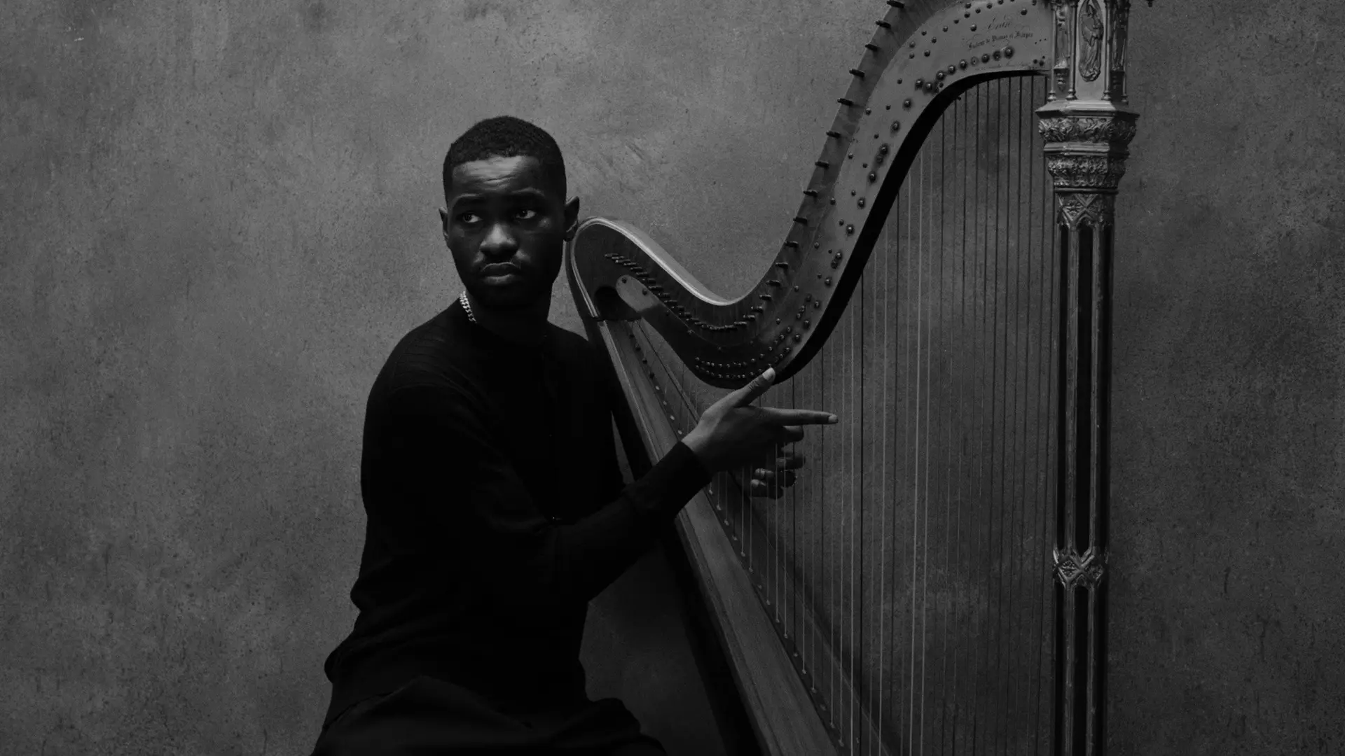 Zwart-witfoto van rapper Dave die naast een grote harp zit en met zijn rechterhand naar de snaren wijst, met een neutrale gezichtsuitdrukking en een effen achtergrond.
