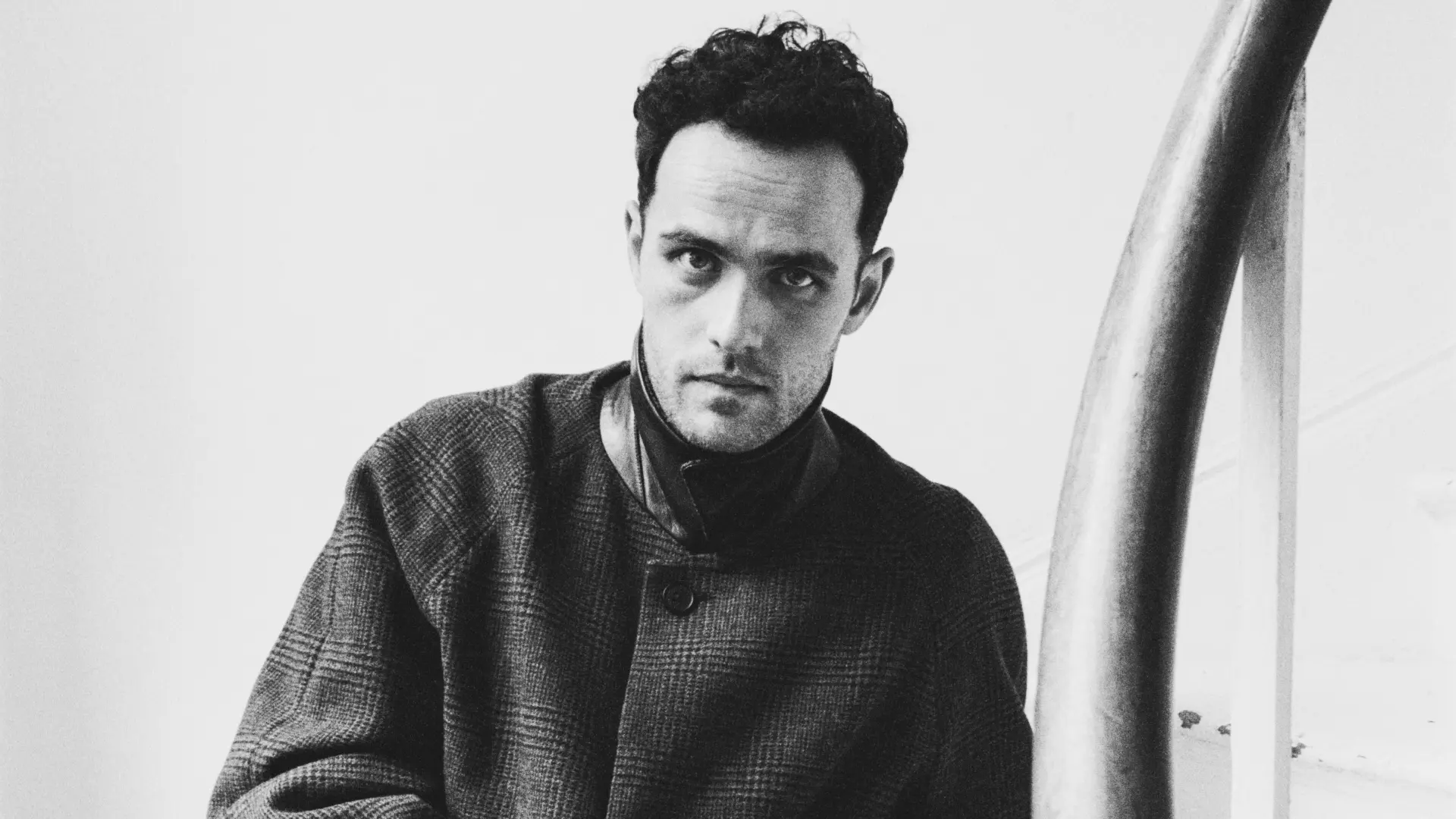 Jordan Rakei zit op een trap in een zwart-witportret en kijkt recht in de camera, gekleed in een lange jas en een geruite broek.