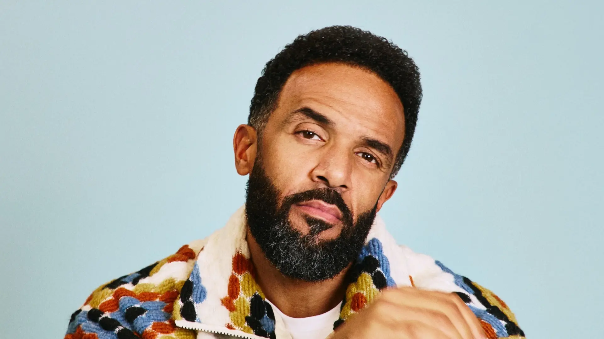 Craig David poseert met gekruiste armen in een crèmekleurige jas met kleurrijke patronen, tegen een rustige lichtblauwe achtergrond.