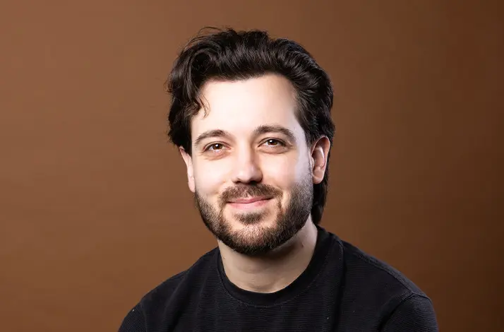 Portret van Reuben Solo met donker, licht golvend haar en korte baard, glimlachend naar de camera. Hij draagt een zwart T-shirt en zit ontspannen met gevouwen handen tegen een warme bruine achtergrond.