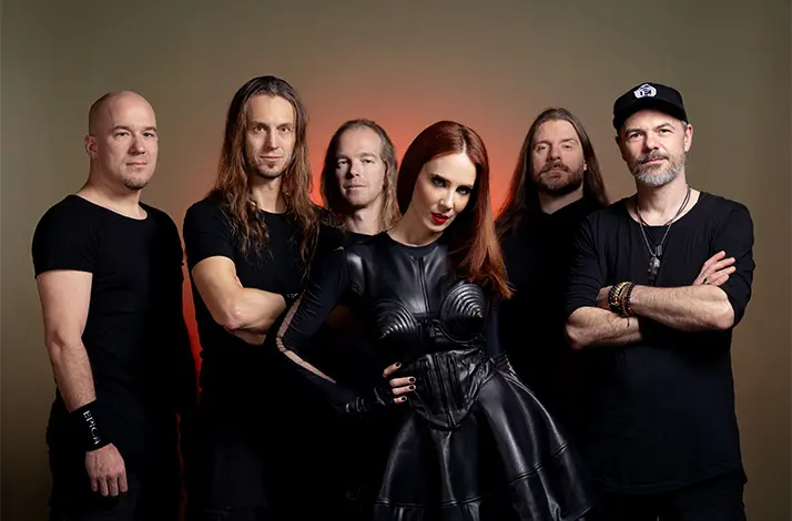 Zes leden van de metalband Epica poseren samen; zangeres centraal in zwarte outfit, bandleden eromheen in zwart, met warme roodachtige achtergrond en studioverlichting.