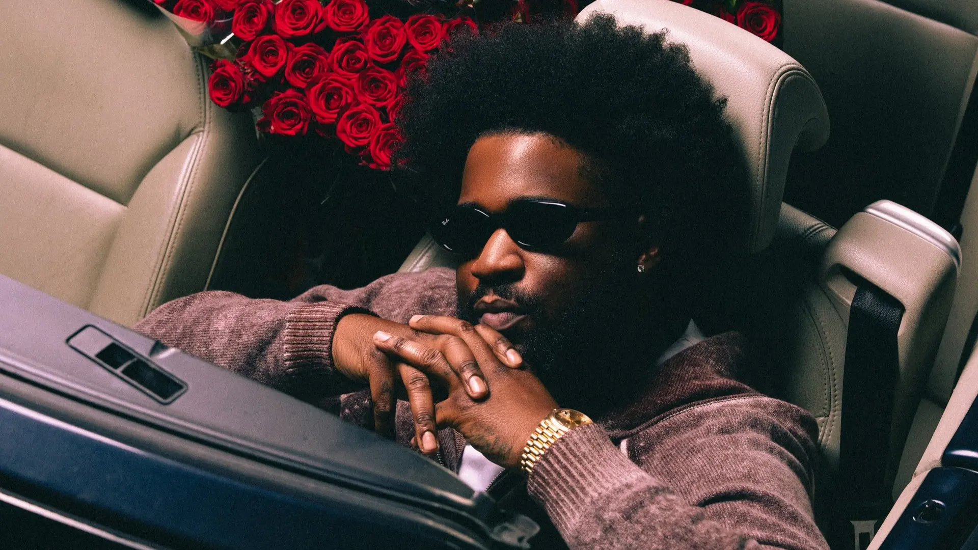 Man met zonnebril zit in een open auto, met op de achterbank meerdere boeketten rode rozen op beige leren stoelen.