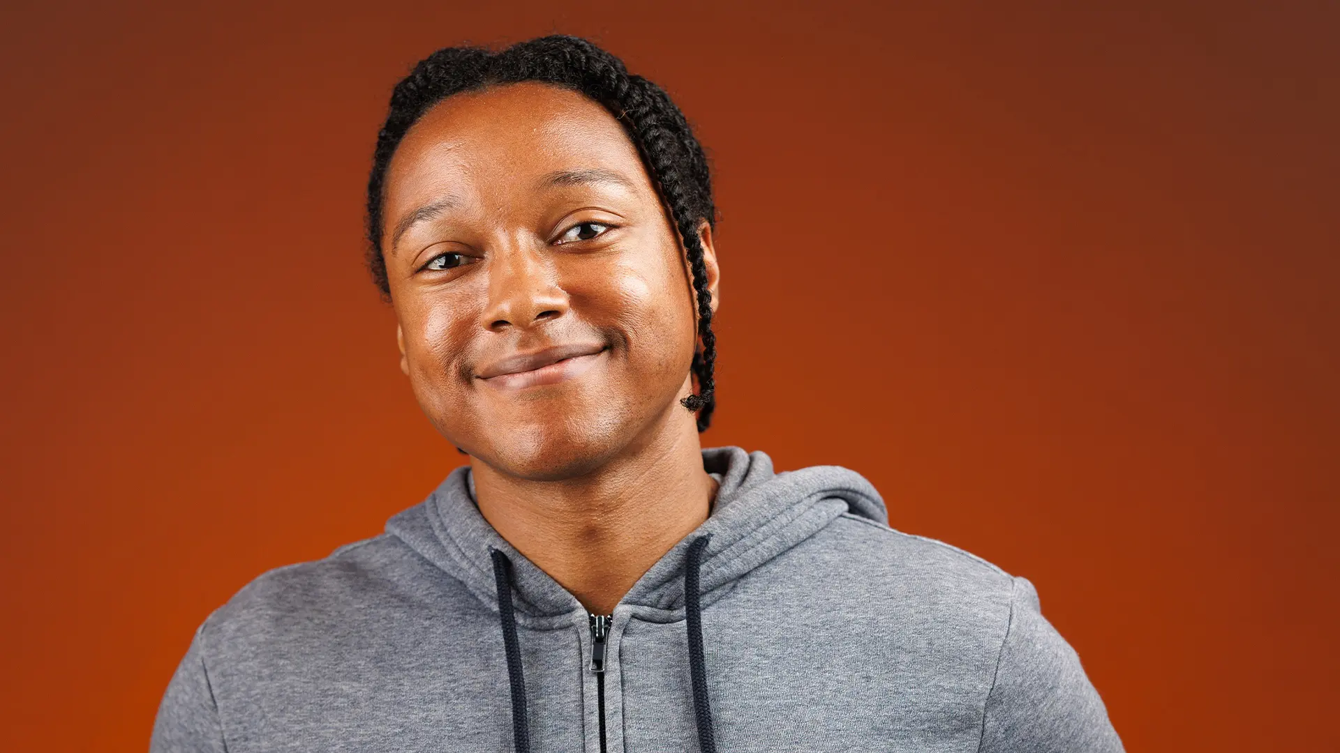 Portret van Josh Johnson met gevlochten haar, lichte glimlach en grijze hoodie met rits en capuchon, handen in de zakken, tegen een effen oranje achtergrond.