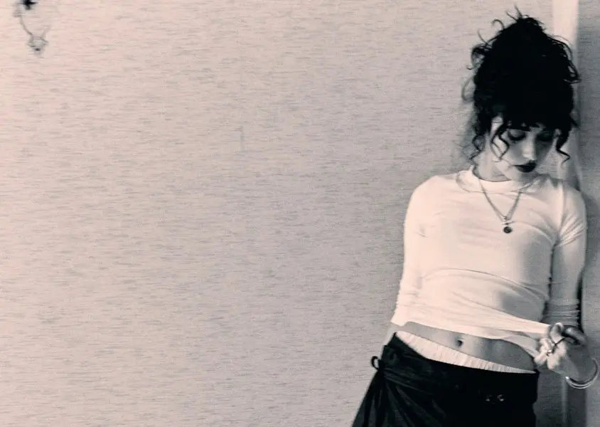 Asha Banks leaning against a wall wearing a white long sleeve top