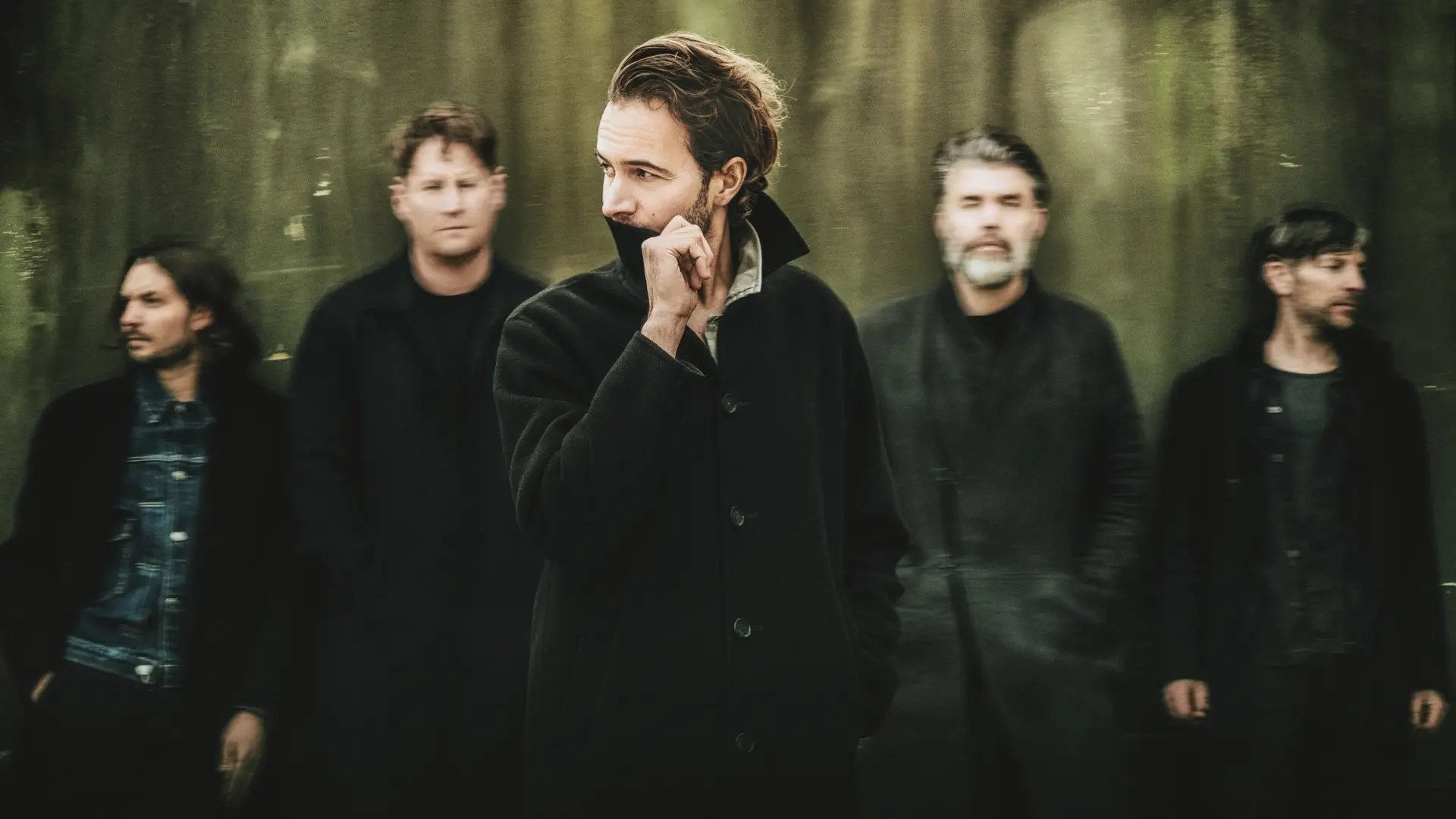 De band Editors poseert in een bosrijke omgeving; de zanger staat scherp in het midden terwijl de overige bandleden wazig op de achtergrond zichtbaar zijn.