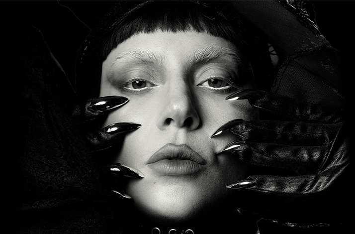 A black and white photo of Lady Gaga's face close to the camera. 2 hands with black gloves are holding her face