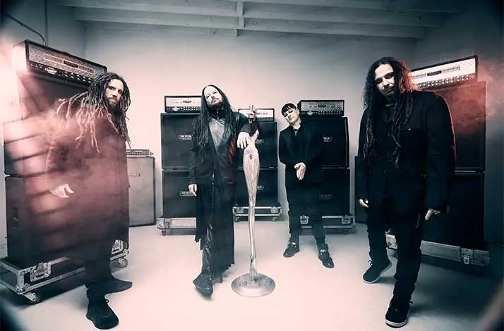 Bandfoto van de metalband Korn. Vier leden in donkere kleding staan in een studio tussen gitaarversterkers; één leunt op een microfoonstandaard, met rook en dramatische belichting.