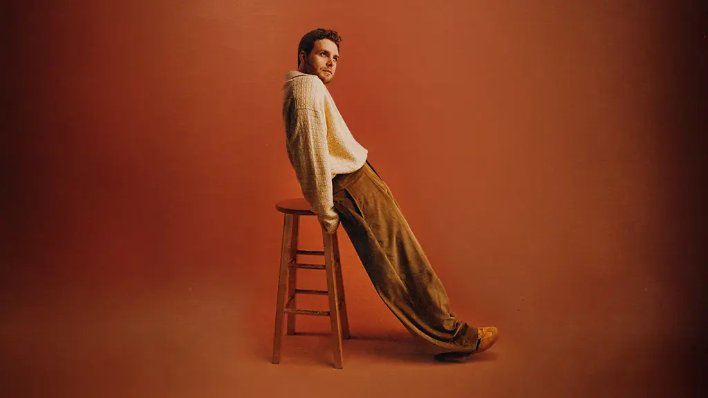 Alex Warren leunt achterover tegen een houten kruk, draagt een lichte trui en wijde bruine broek, tegen een warme oranje achtergrond