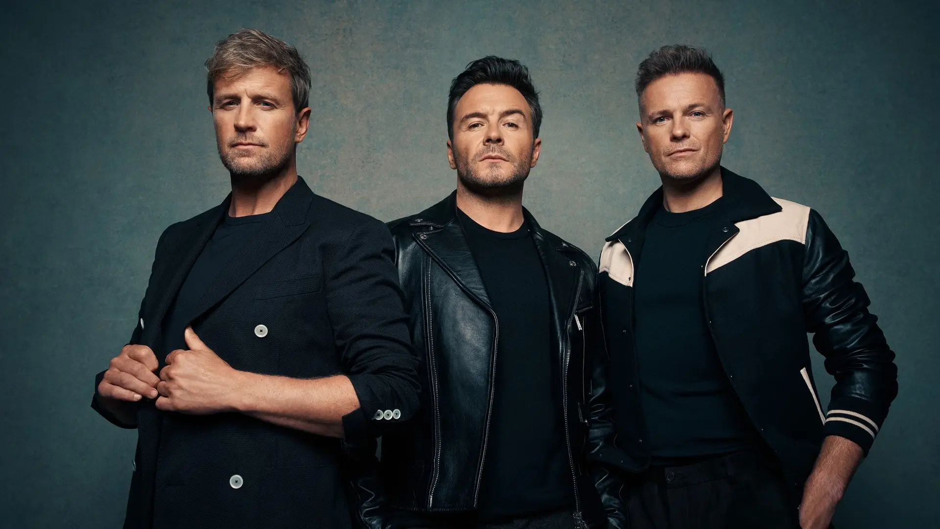 Drie mannen van de Ierse band Westlife poseren voor een effen blauwe achtergrond, gekleed in donkere, stijlvolle outfits; de man links draagt een donkerblazer, de man in het midden een zwarte leren jas en de man rechts een zwart jack met beige details.
