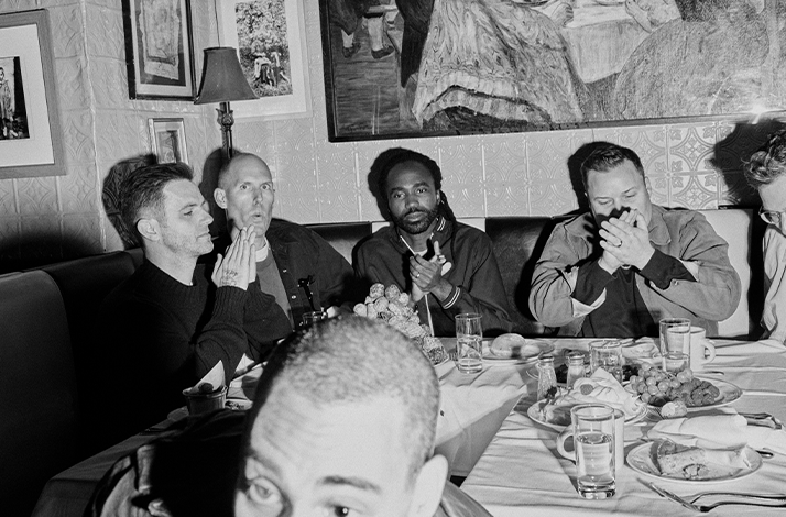 Zwart-witfoto van Bleachers rond een restauranttafel met brood en drank; sommigen lachen of houden handen voor gezicht, informele en gezellige sfeer met kunst aan de muur.