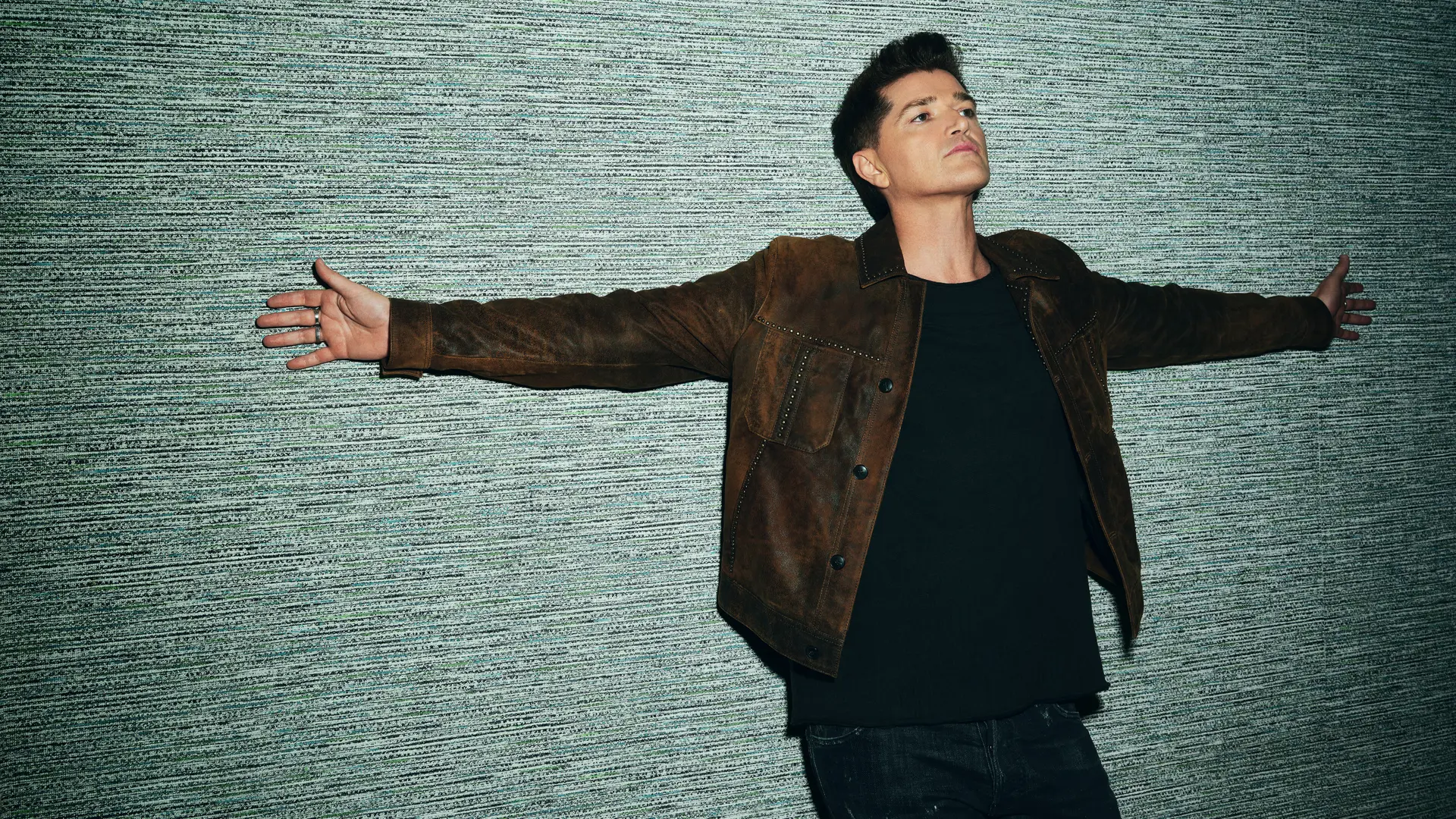 Danny O’Donoghue staat met gespreide armen tegen een grijze muur en kijkt omhoog terwijl hij een donkerbruin suède jack draagt