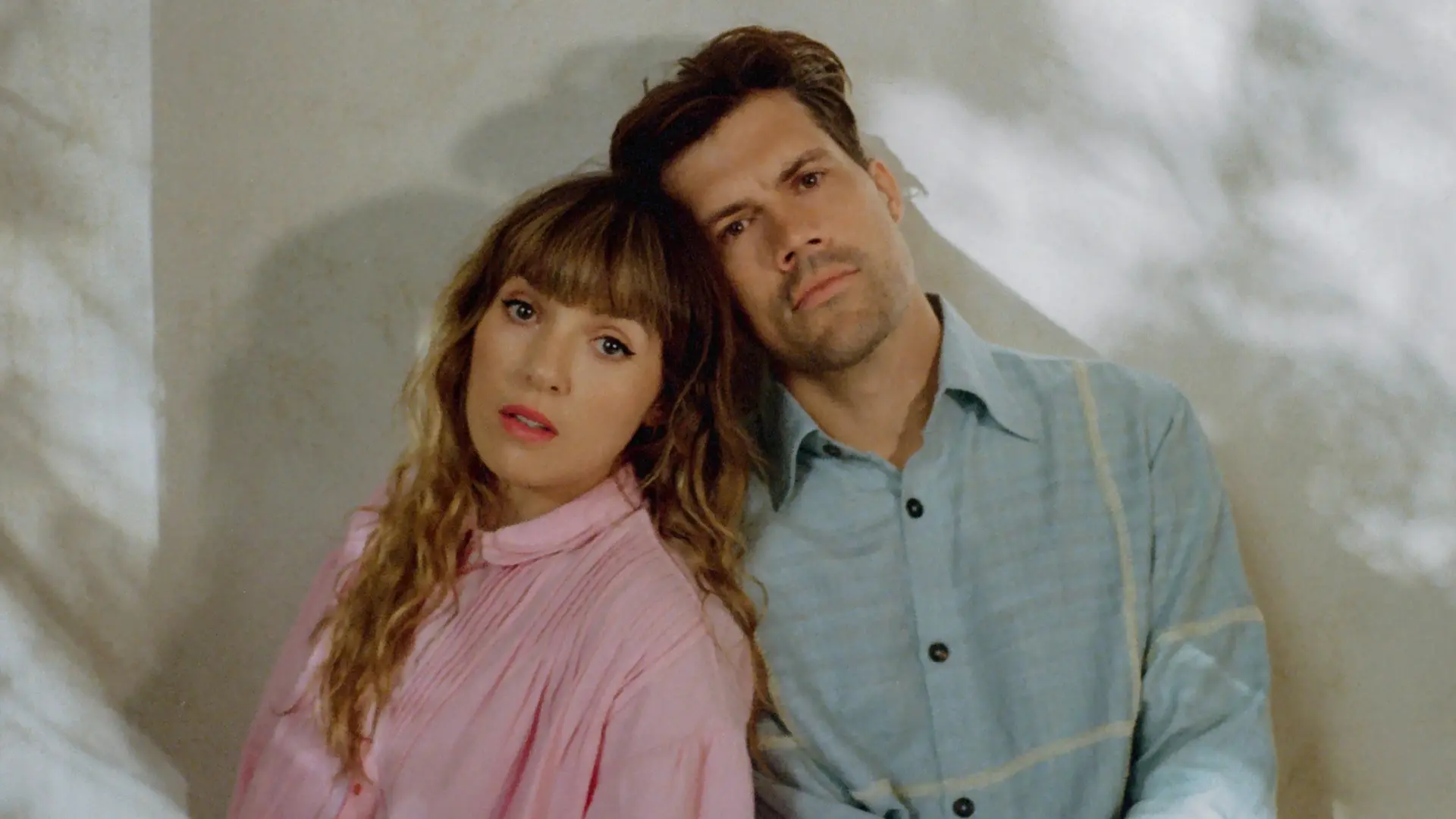 Het duo Oh Wonder leunt met de hoofden tegen elkaar en kijkt in de camera, gekleed in een roze blouse en lichtblauw overhemd, tegen een lichte muur met zachte schaduwen.