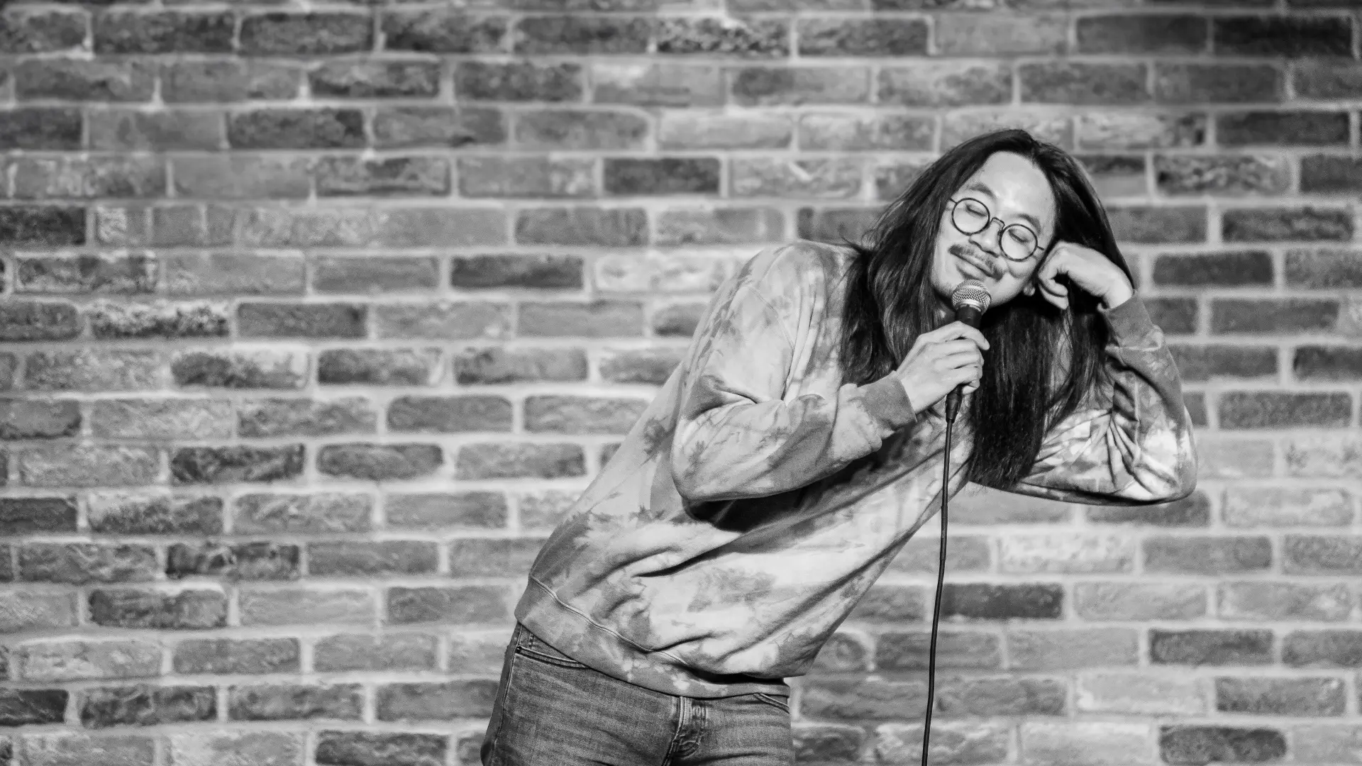 Zwart-witfoto van Sheng Wang op een podium voor een bakstenen muur; hij houdt een microfoon vast, leunt met zijn hoofd op zijn hand en glimlacht met gesloten ogen.