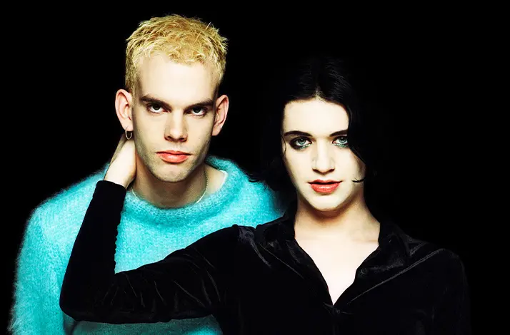 Twee leden van Placebo poseren tegen zwarte achtergrond; links één met blond haar en turquoise trui, rechts de ander met zwart haar en donkere blouse, intens kijkend, hand rust op schouder.