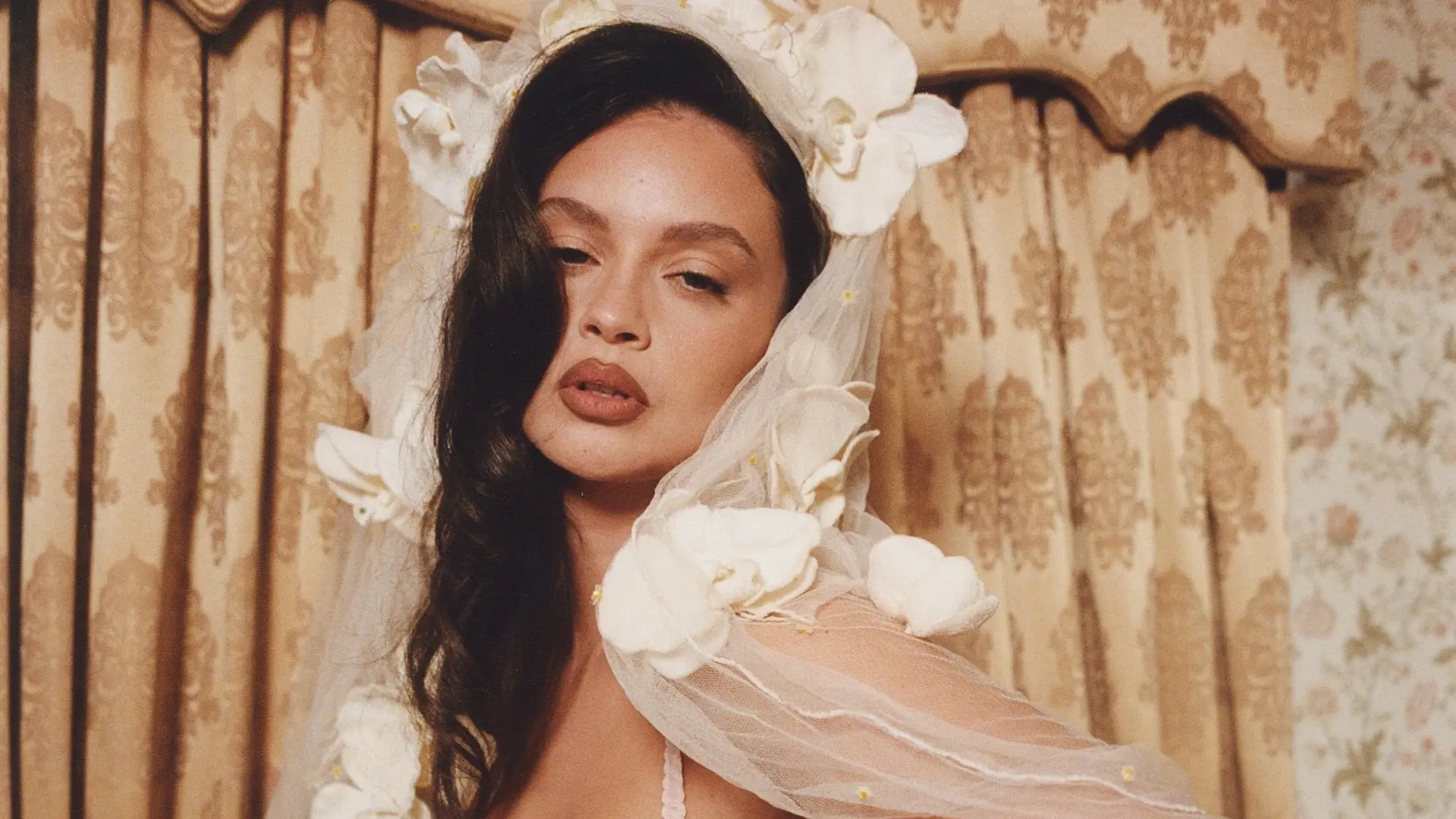 Portret van Sabrina Claudio met lange, golvende donkere haren, gekleed in een doorschijnende sluier versierd met grote witte bloemen. Op de achtergrond beige gordijnen met een barok patroon.