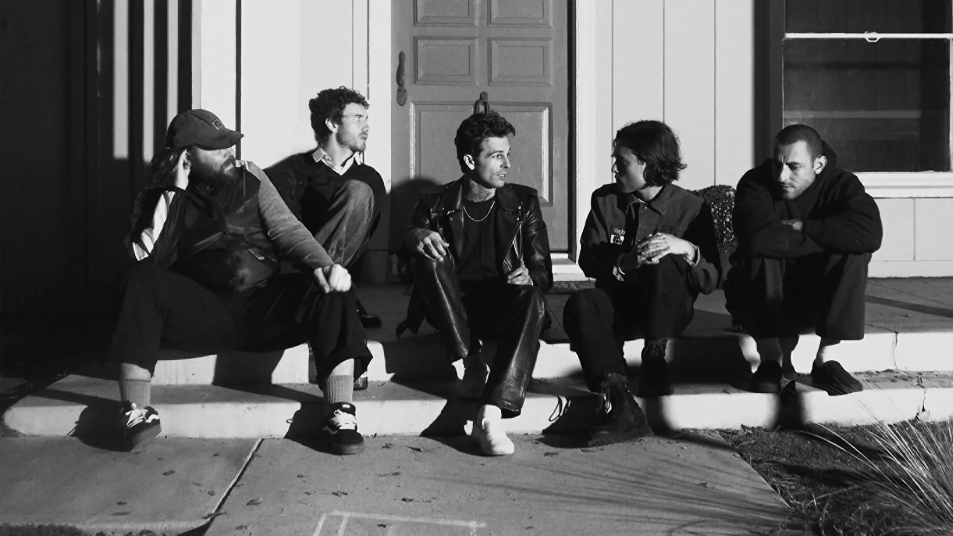 Zwart-witfoto van vijf mannen die op de stoep voor een huis zitten, met een gesloten deur en buitenlamp achter hen. Op het trottoir is met krijt een pijl en het woord “LOVE” getekend.