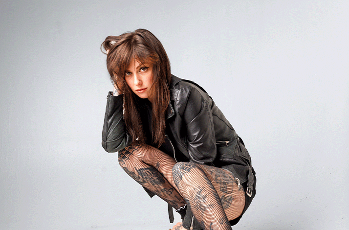 Natalie Cuomo met lang donker haar hurkt in een lichte studio. Ze draagt een zwarte leren jas, visnetpanty’s met tatoeages zichtbaar op haar benen en zwarte platformschoenen met een ster op de zijkant. Ze kijkt recht in de camera.