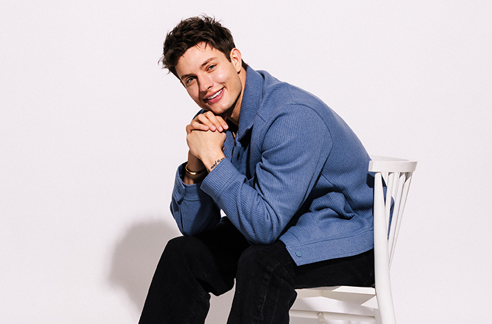 Matt Rife sitting on a chair in front of a white wall, he's smiling