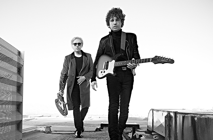 A black and white photo of The Kooks walking on a seaside boulevard, holding guitars