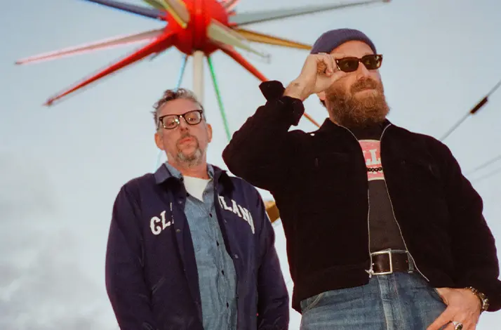 Twee leden van de band The Black Keys staan buiten gefotografeerd vanuit een laag standpunt, met een kleurrijke stervormige lichtsculptuur achter hen. Ze dragen casual kleding; één man draagt een bril, de ander een baard en zonnebril.