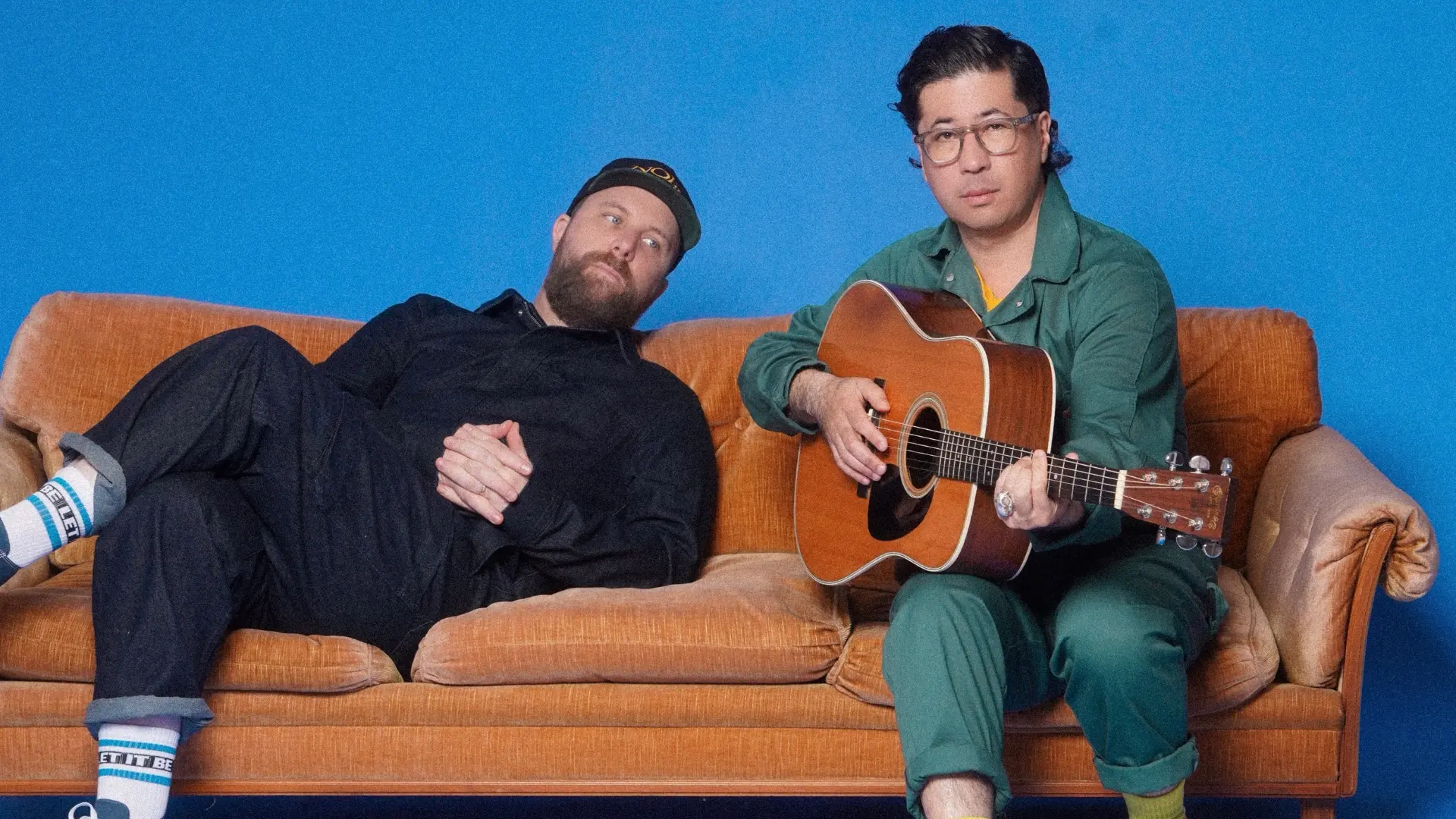 Zwei Mitglieder von Choir!Choir!Choir!, die auf einer orangenen Couch sitzen. Der linke lehnt gemütlich drauf, der rechte hat eine Gitarre in der Hand. Beide schauen in die Kamera. Der Hintergrund ist tiefblau