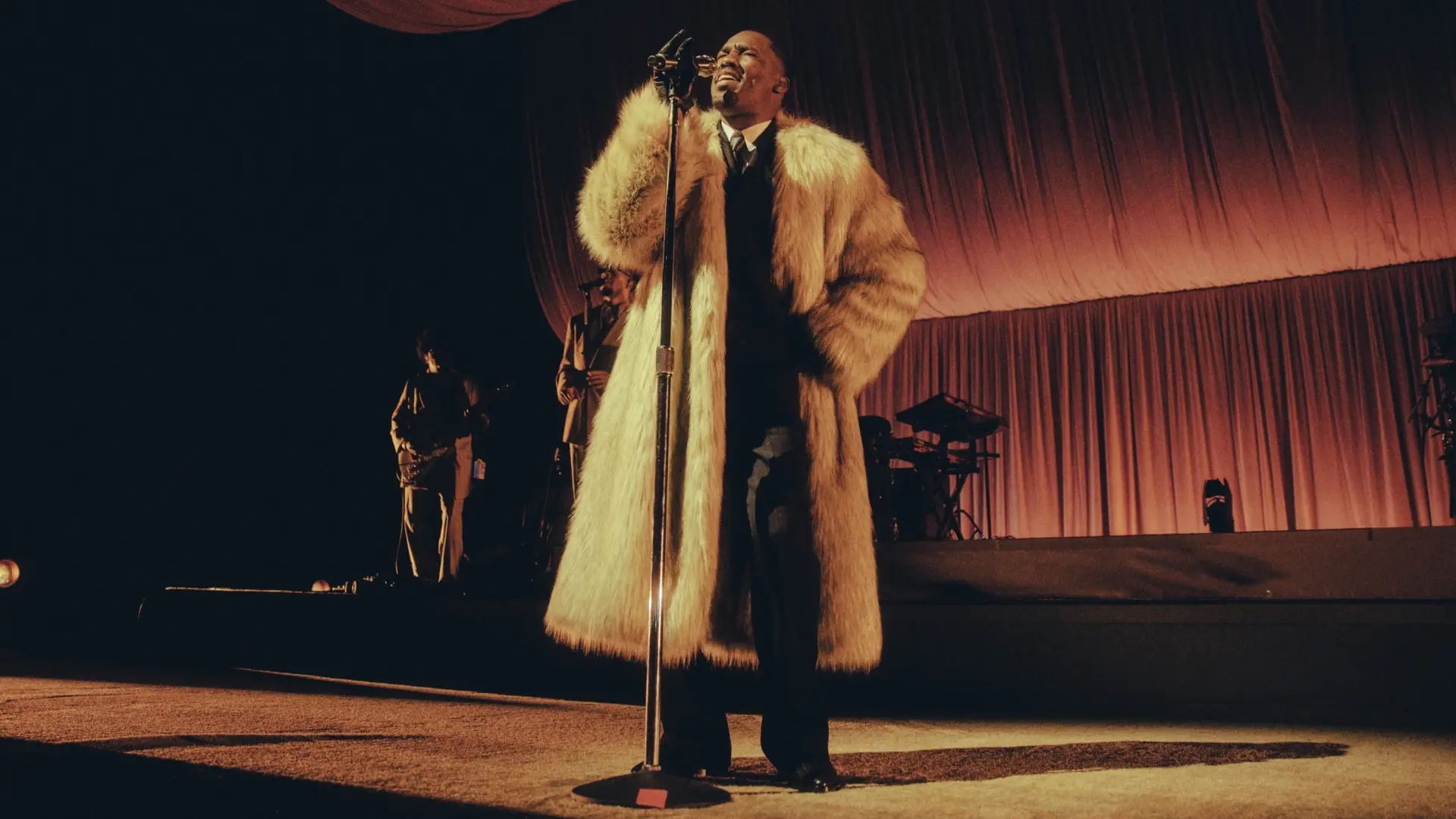 Zanger Giveon op een podium, gekleed in een lange bruine bontjas, zingend in een microfoon met een warm oranje verlichte achtergrondgordijn en muzikanten op de achtergrond.