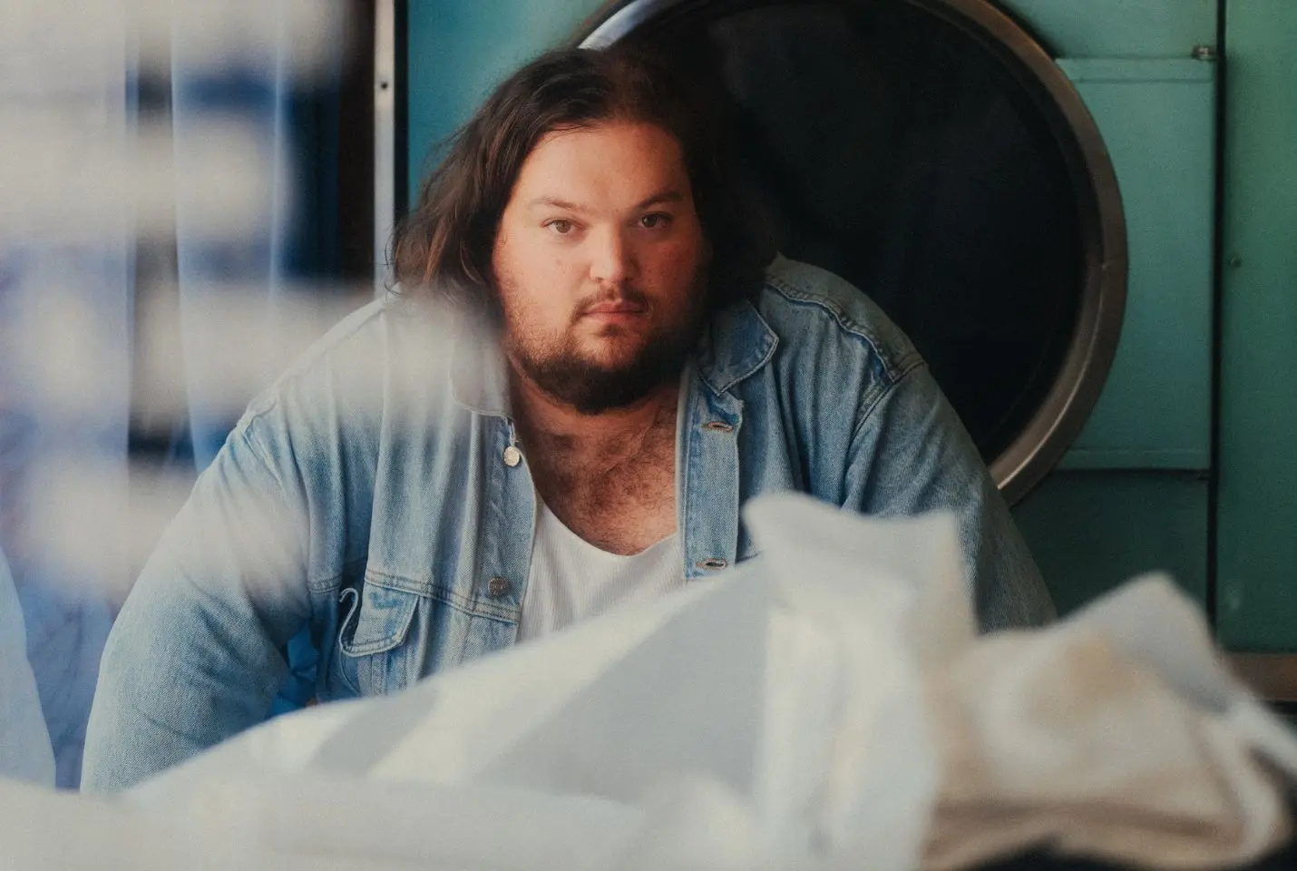A man with dark hair and a beard wearing a white tshirt and denim shirt
