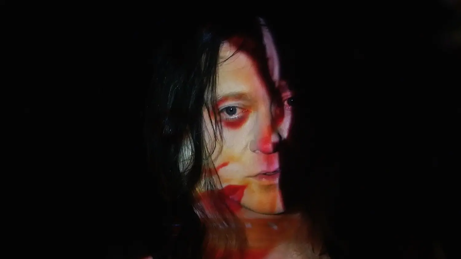 Portret van ANOHNI, half zichtbaar gezicht met nat donker haar, belicht met rood en oranje licht tegen een zwarte achtergrond.