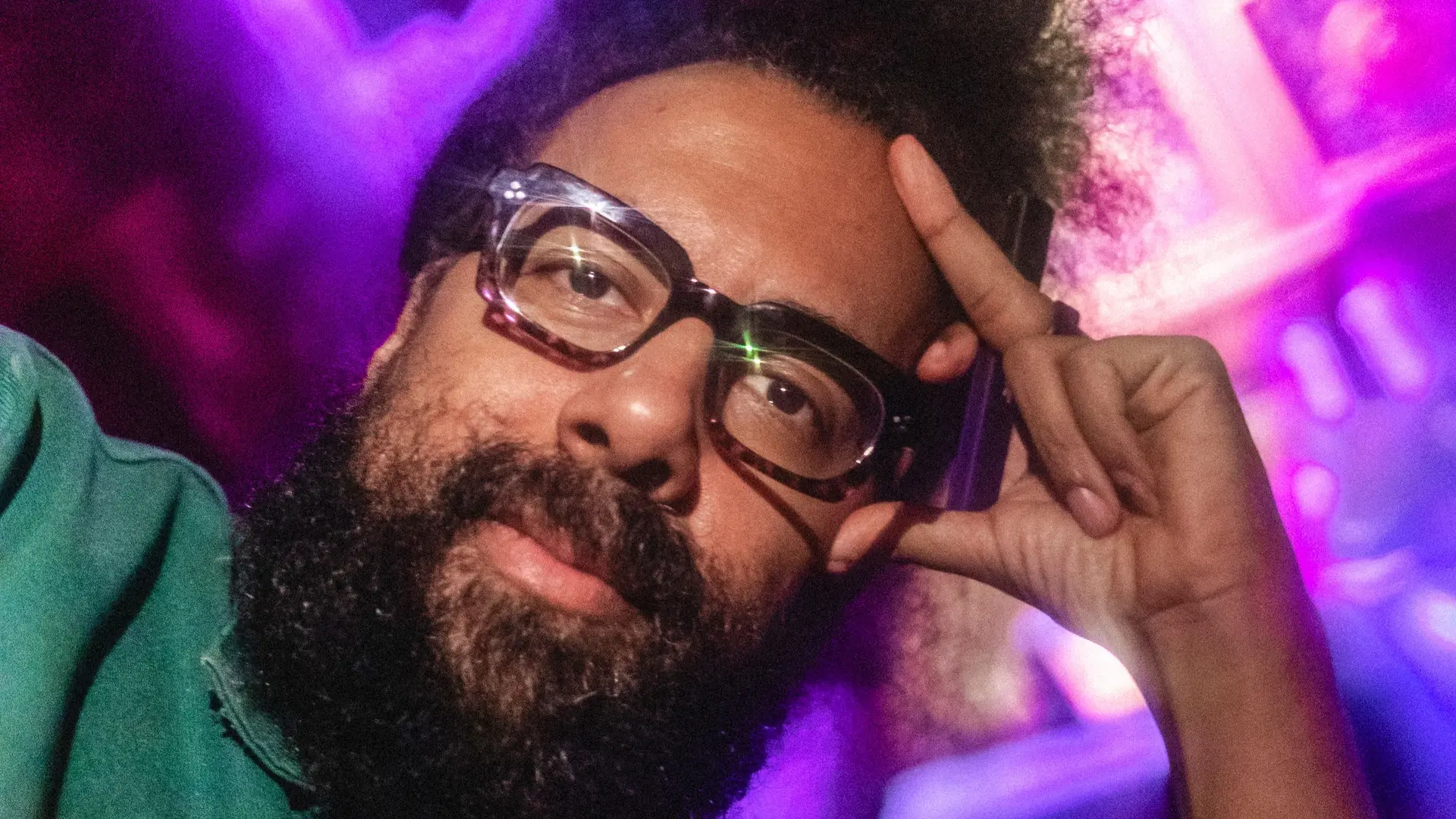 Reggie Watts met bril, baard en hoog opgestoken haar in een groen overhemd, gefotografeerd tegen een paars-roze verlichte achtergrond met een artistieke, intieme sfeer.