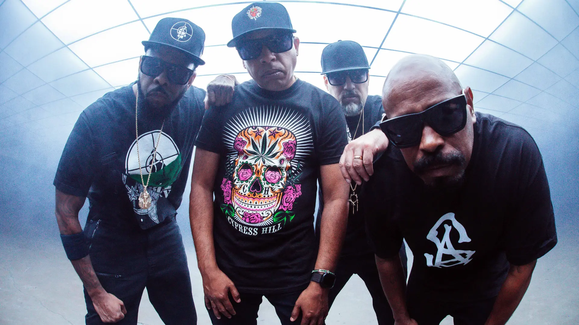 Vier mannen in donkere kleding, met petten en zonnebrillen, kijken recht in de camera. Ze staan in een lichte, futuristische ruimte.