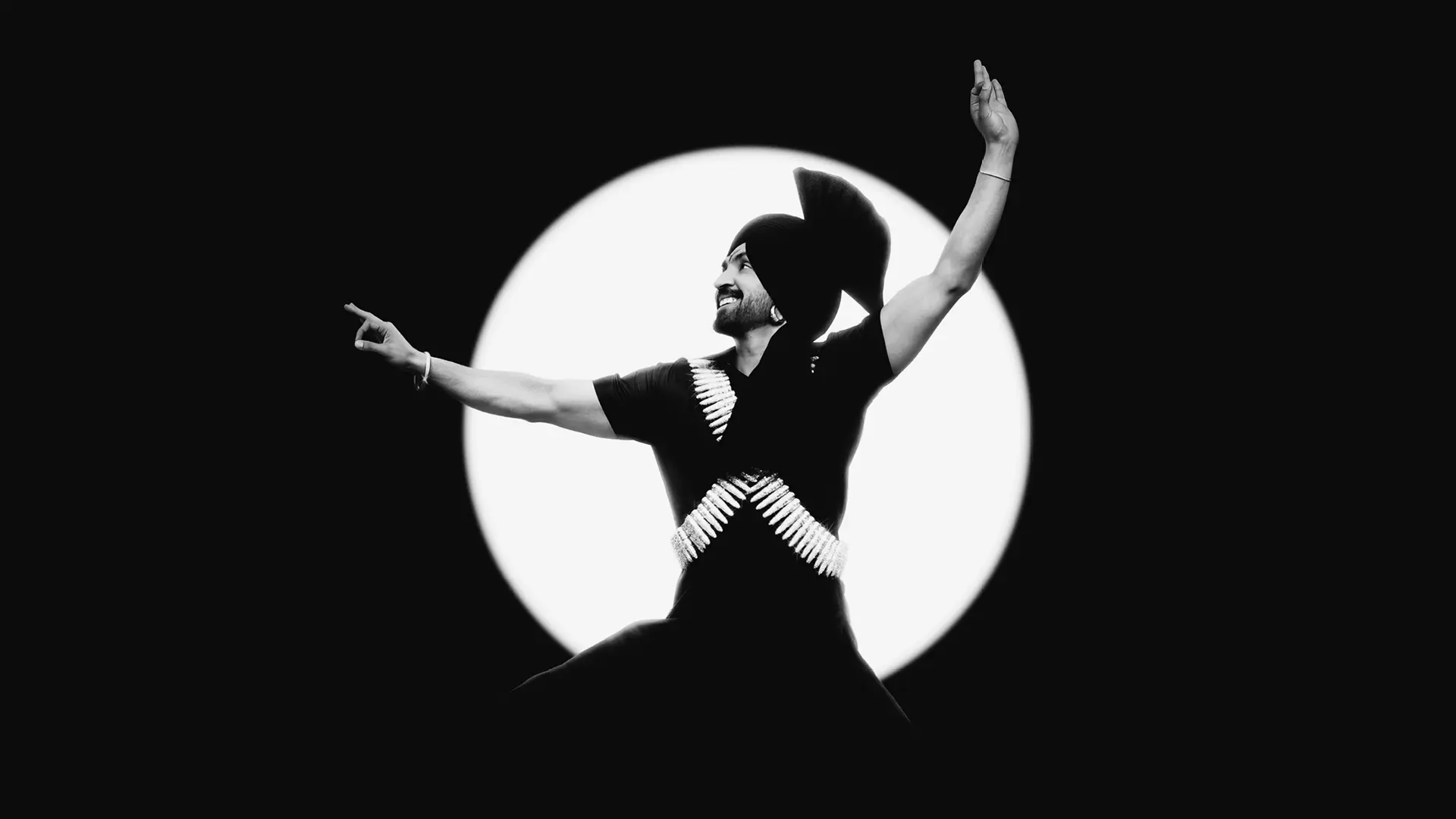 Diljit Dosanjh poseert dansend met uitgestrekte armen voor een grote ronde lichtbron, afgebeeld in zwart-wit tegen een donkere achtergrond.