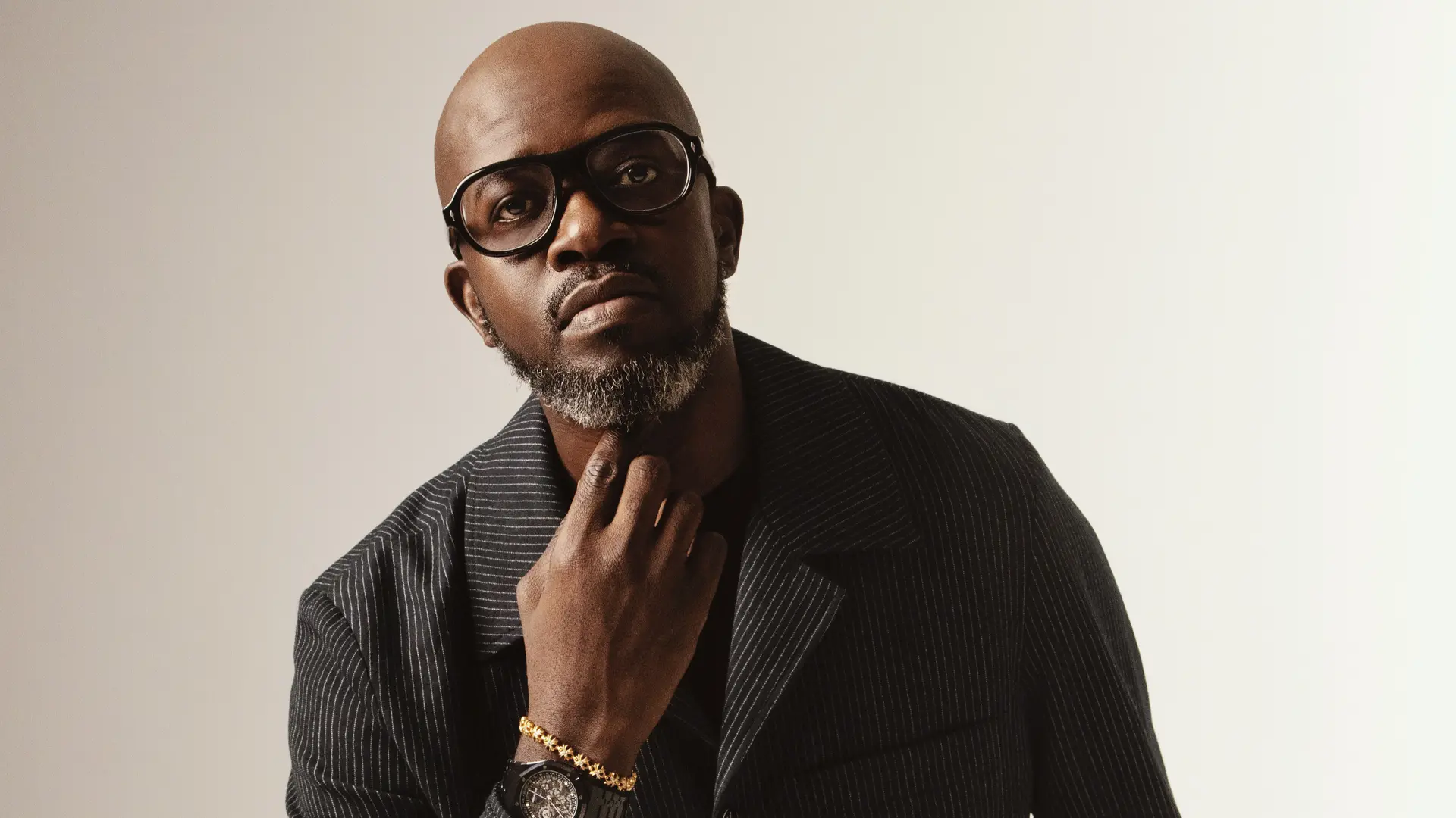 Black Coffee zit op een stoel in een donkere outfit en bril, met zijn hand onder zijn kin, tegen een rustige lichte achtergrond.