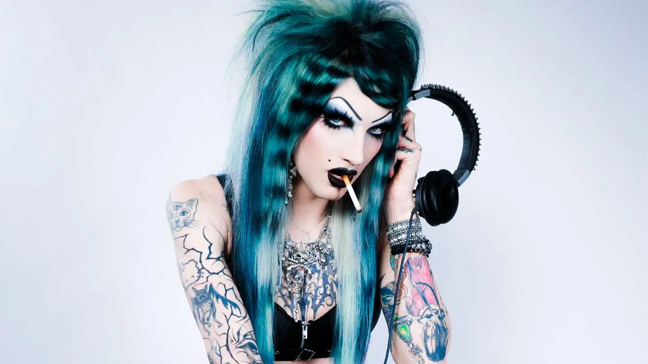 "Person with teal hair, bold makeup, tattoos, and black clothing holding headphones with a cigarette in mouth."