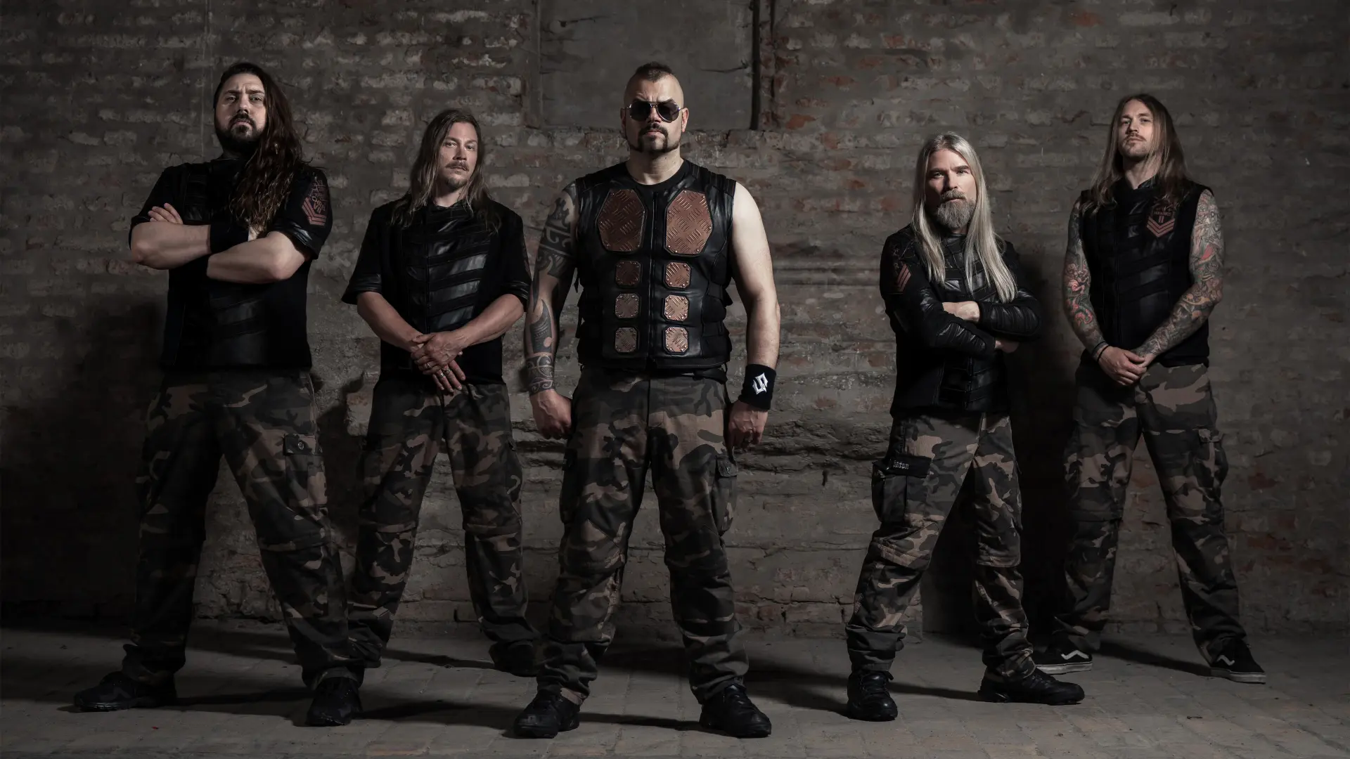 Leden van Sabaton staan naast elkaar in een industriële ruimte met bakstenen muur, gekleed in zwarte vesten en camouflagebroeken. Eén persoon draagt een zonnebril en borstpantser, twee personen hebben zichtbare tatoeages.