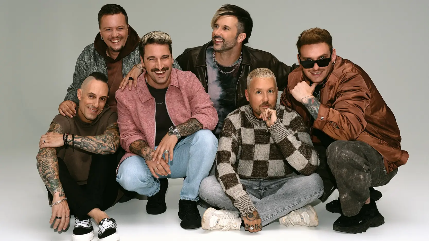 Groepsfoto van zes lachende mannen van Electric Callboy in casual kleding, met zichtbare tatoeages en verschillende moderne kapsels; drie zitten op de grond, drie poseren erachter tegen een neutrale achtergrond.