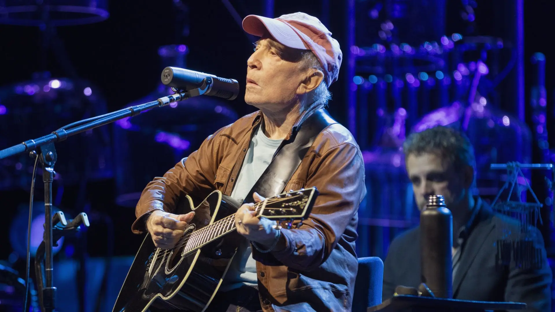 Paul Simon zittend op een podium, zingend in een microfoon en spelend op een akoestische gitaar, met pet en bruine jas, tegen een blauw verlichte achtergrond.