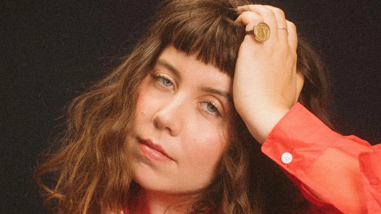A woman with wavy brown hair and bangs gazes softly at the camera, resting her hand against her forehead. She wears a bright red blouse and a gold ring, with a dark background behind her.
