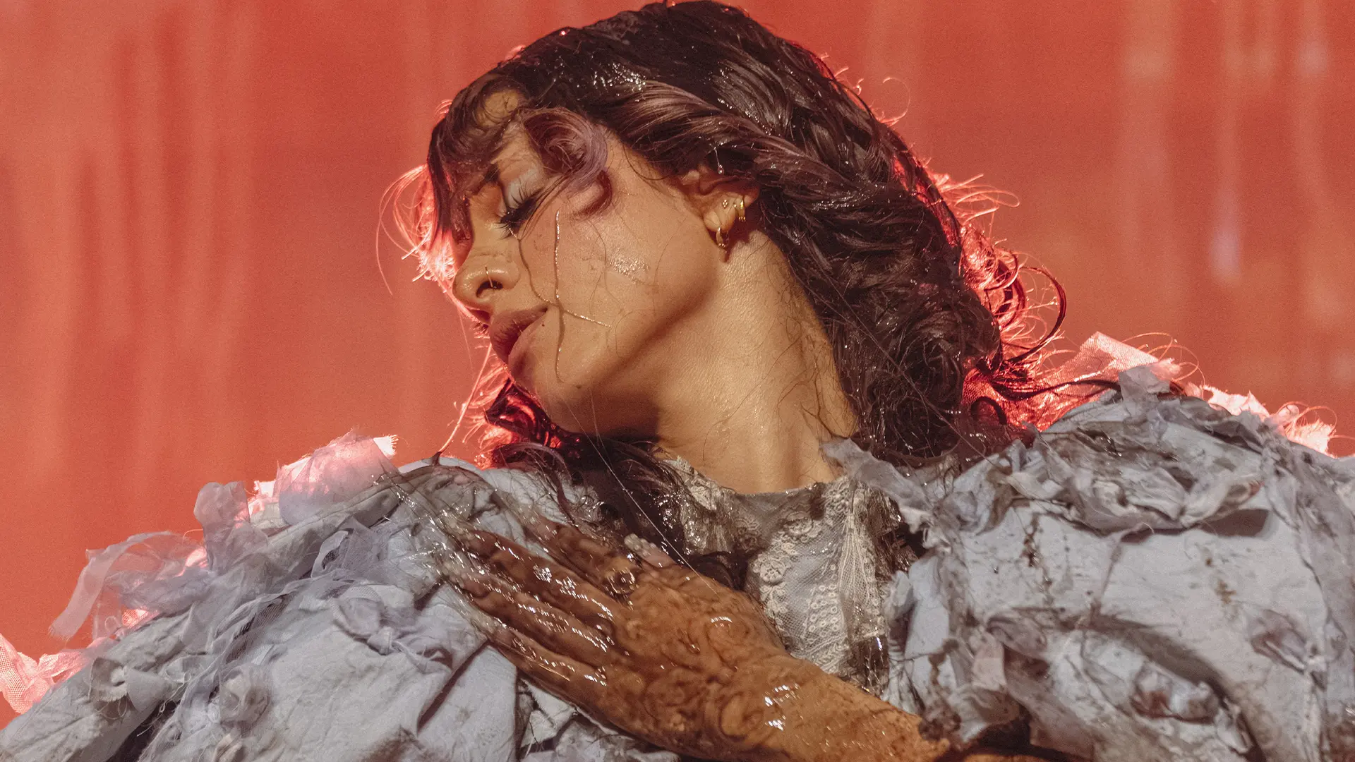 Portret van Melanie Martinez met nat haar en waterdruppels op haar gezicht, hoofd naar achteren gedraaid en ogen gesloten, met een grijze outfit met textuur en een natte hand op haar schouder tegen een warme oranje achtergrond.