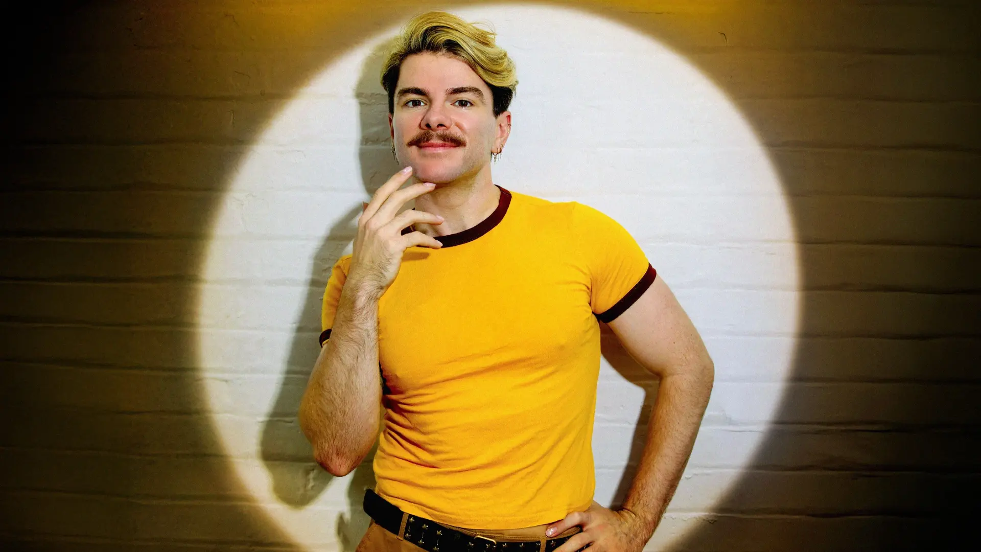 Christopher Hall staand voor een witte bakstenen muur, belicht door een ronde spotlight, gekleed in een geel T-shirt met donkerrode randen en beige broek, met één hand bij zijn gezicht en de andere in zijn zij.
