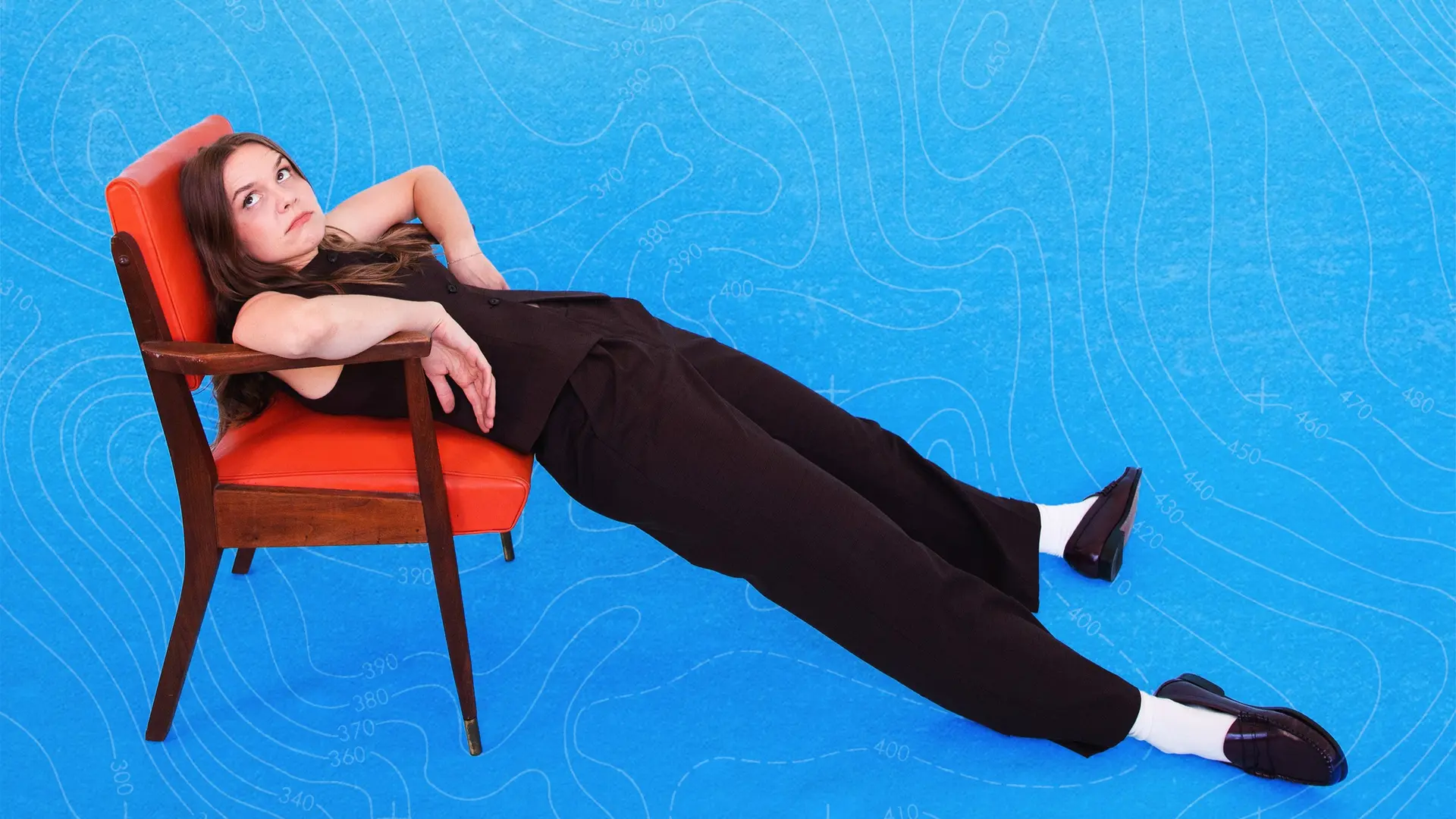 Comedian Laura Ramoso ligt half onderuitgezakt in een vintage oranje stoel, met haar benen languit op een felblauwe achtergrond met subtiele lijnpatronen. Ze kijkt licht ironisch omhoog, gekleed in een donkere outfit met witte sokken en zwarte loafers