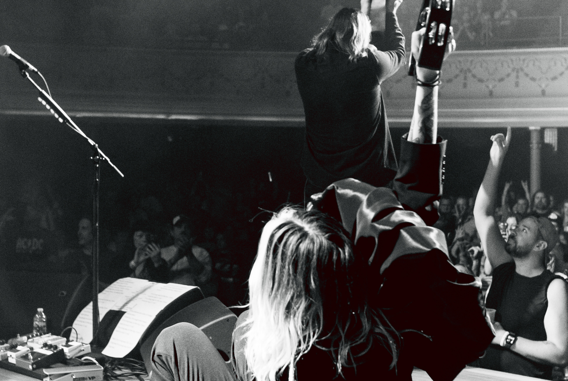 Black and white image of band members of Band of Skulls on stage taken from behind them with the crowd in front with one fan in the bottom right corner with his hand in the air.